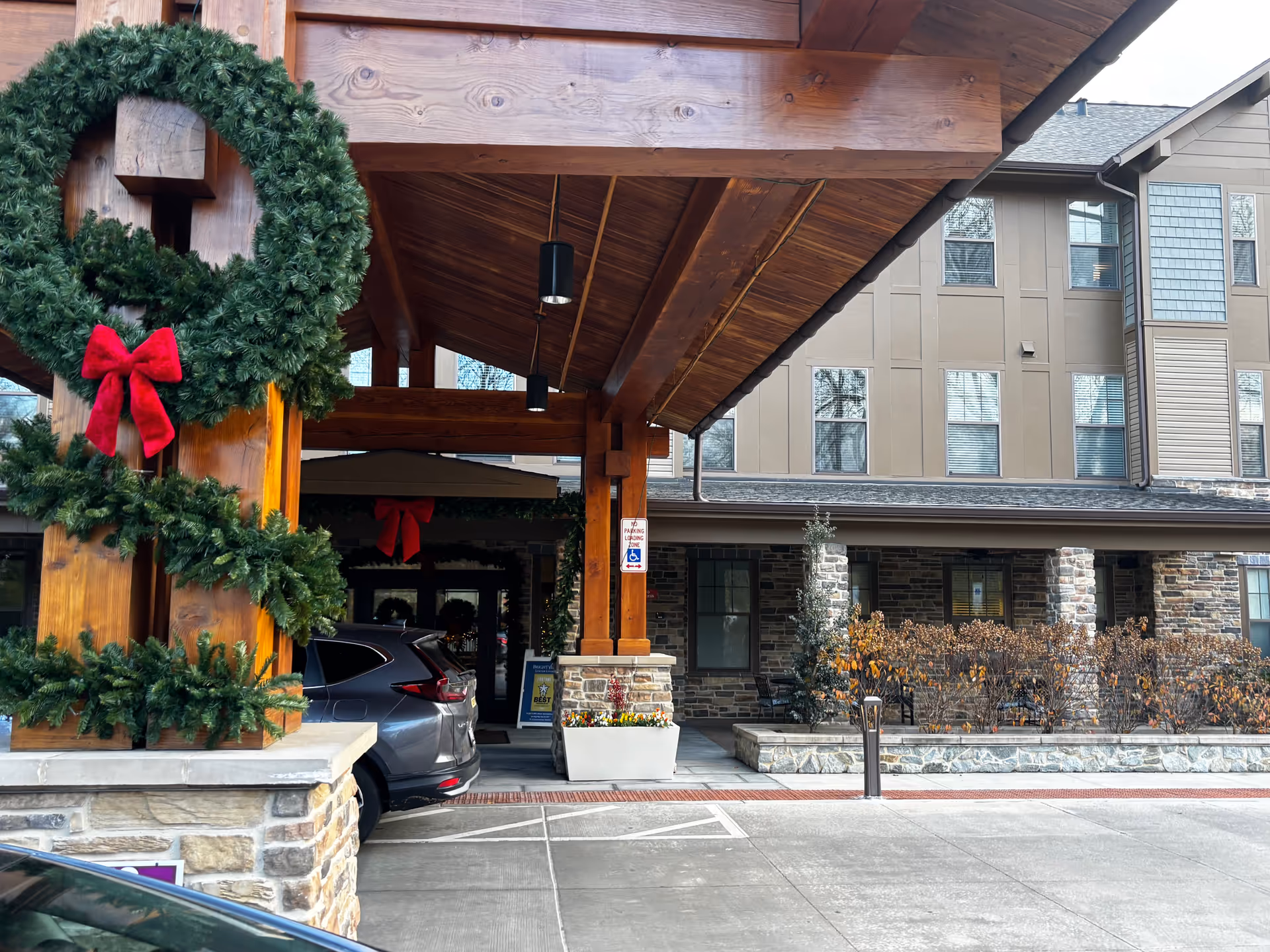 Entrance of a senior living facility decorated with a large green wreath with a red bow on a wooden pillar. The building has stone and beige siding with multiple windows. There is a covered drop-off area with a parked gray SUV and a sign indicating a no parking loading zone for handicapped access. Some plants and bushes are visible near the entrance.