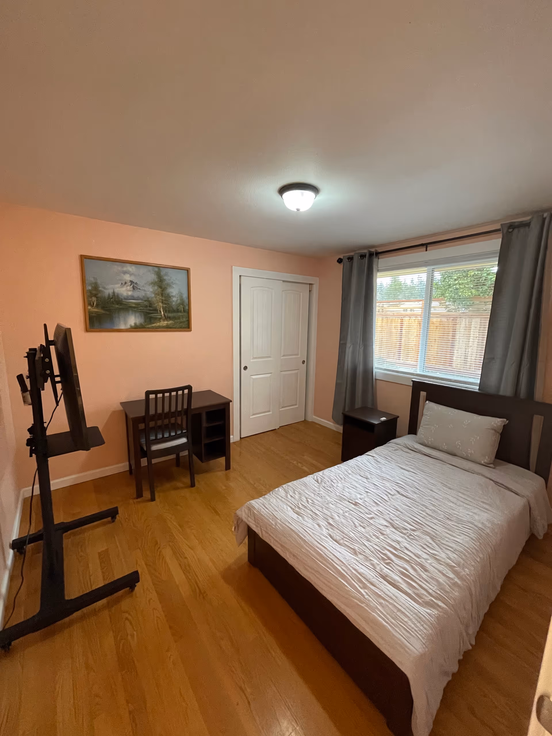 A small bedroom in a senior living facility with a bed, desk, and window.