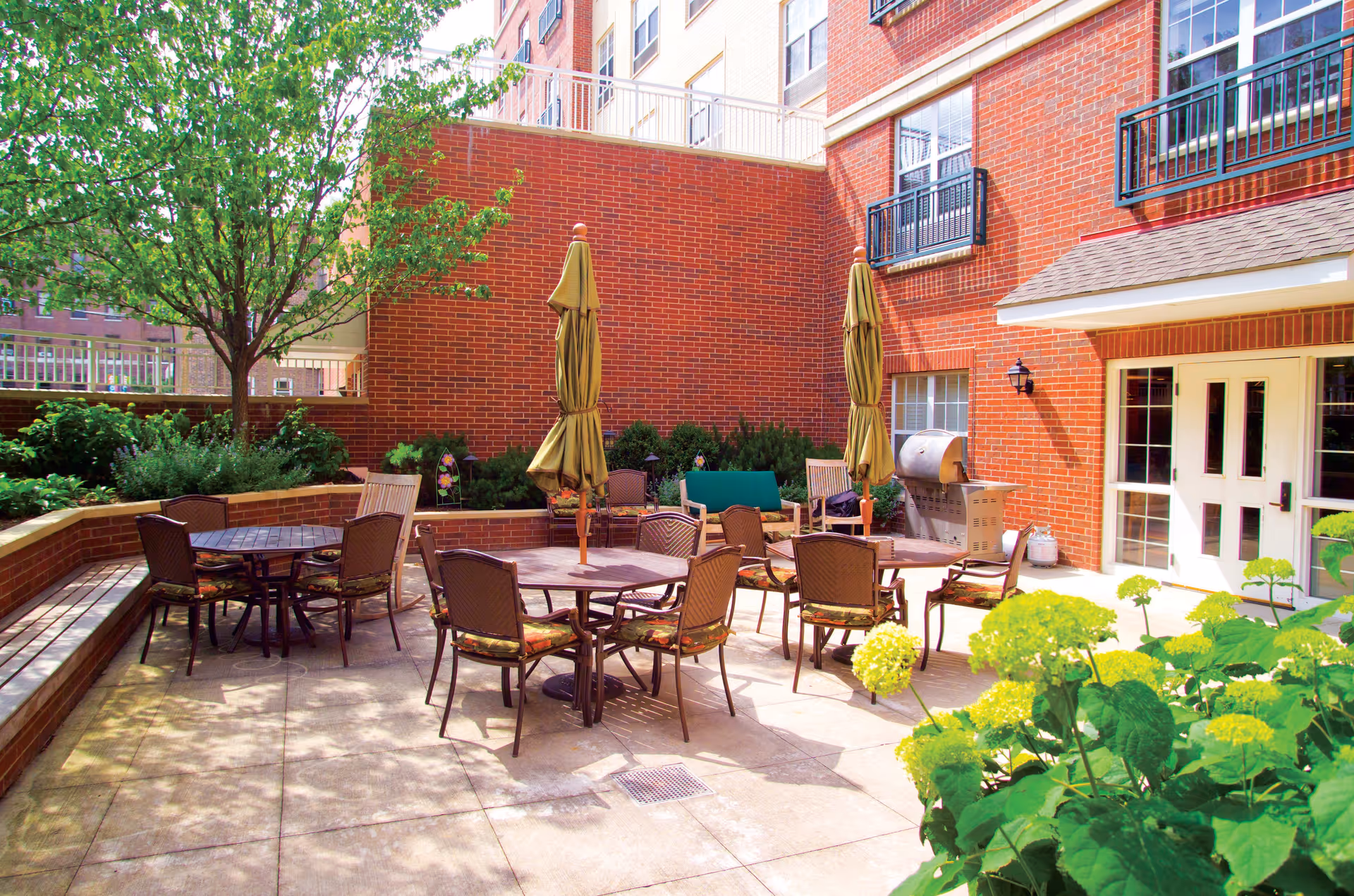 Outdoor patio area at Belmont Village Senior Living Oak Park with several round tables and chairs, two closed umbrellas, a built-in brick bench, green plants, and a barbecue grill against a red brick building.