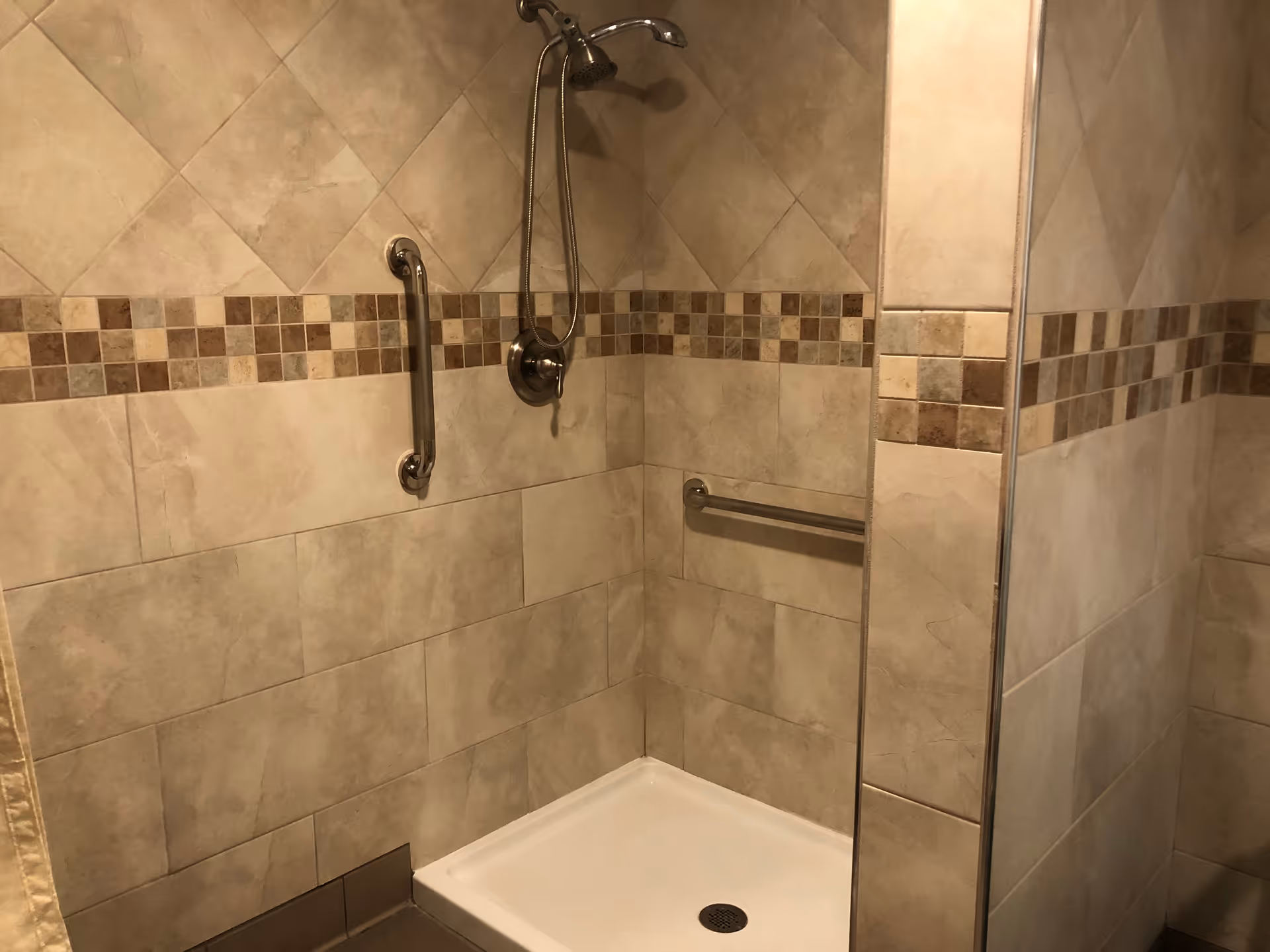 A tiled shower area with beige and brown tiles, featuring a handheld showerhead, a grab bar on the wall, and a small built-in bench or ledge.