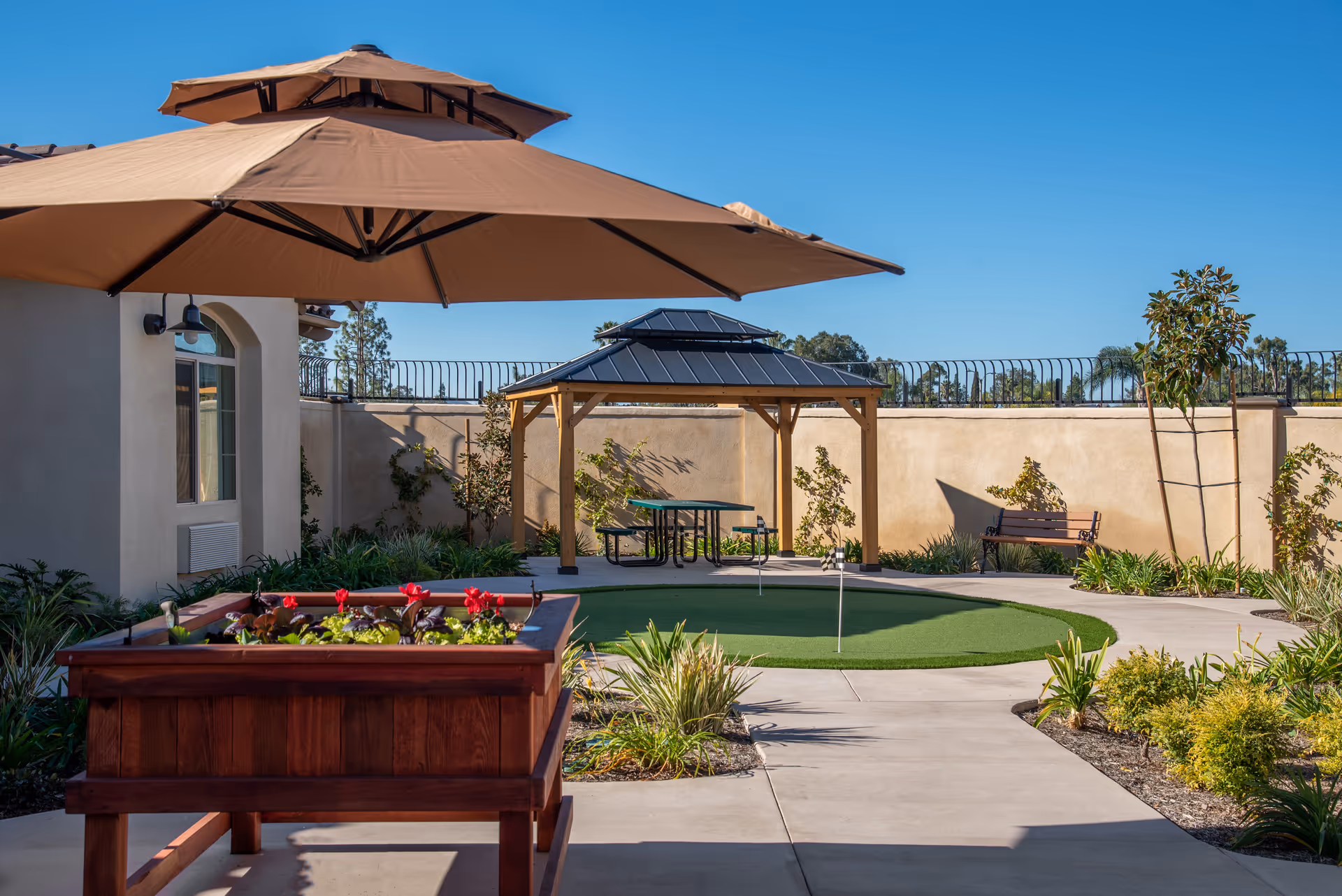 Outdoor courtyard with a gazebo, large patio umbrella, raised planter, seating and a small putting green under a clear blue sky.