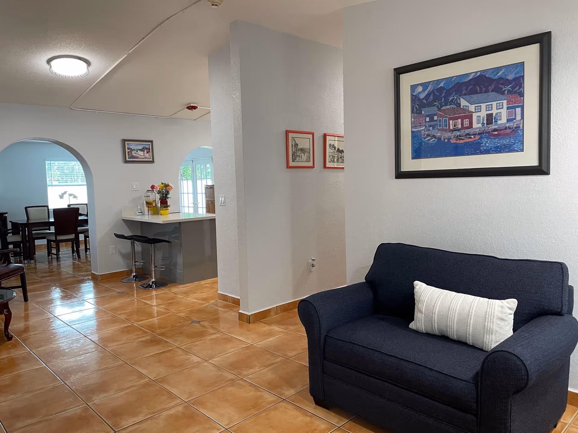 Interior view of a senior living facility showing a navy blue loveseat with a white pillow against a white wall with a framed painting above it. The room has brown tiled flooring and white walls. In the background, there is a kitchen counter with two black bar stools and a dining area with a table and chairs visible through an arched doorway.