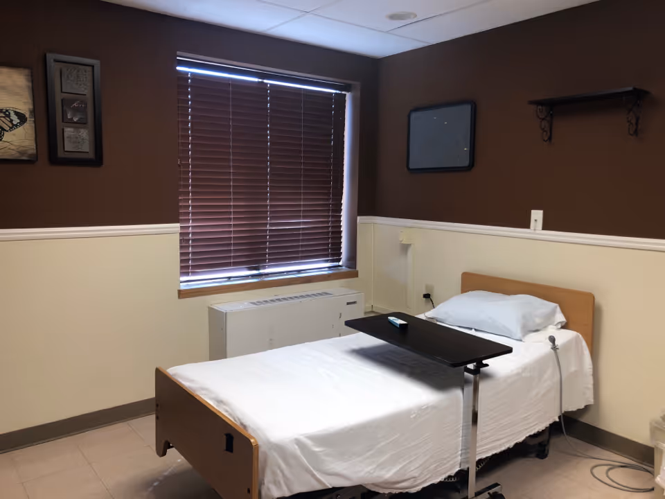 A simple bedroom in a senior facility with a bed, window blinds, and a tray table.