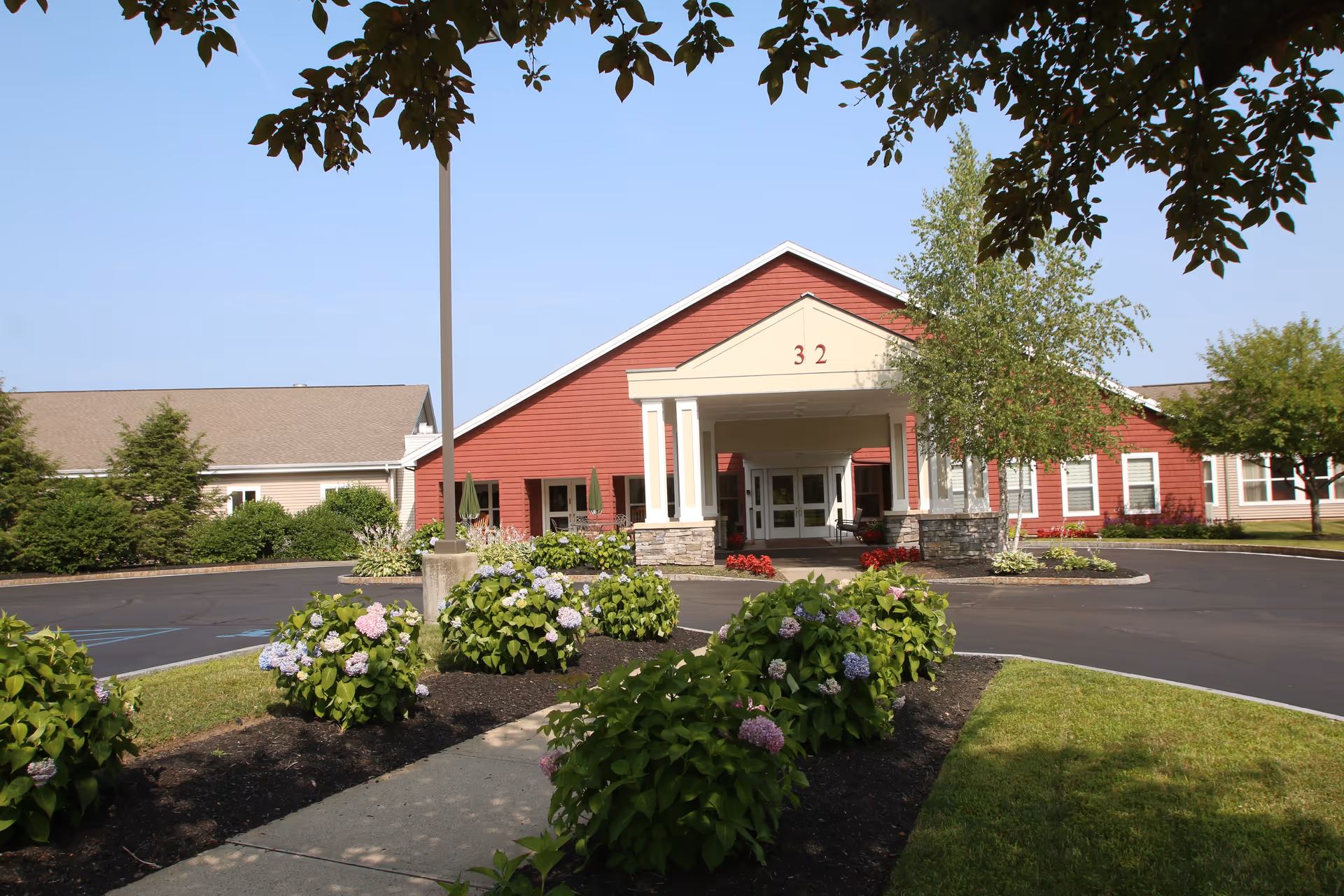 Front exterior view of a senior living facility building with a red facade and white columns at the entrance. The building number 32 is displayed above the entrance. There are landscaped flower beds with blooming hydrangeas and a paved driveway leading to the entrance. Trees and bushes surround the area under a clear blue sky.