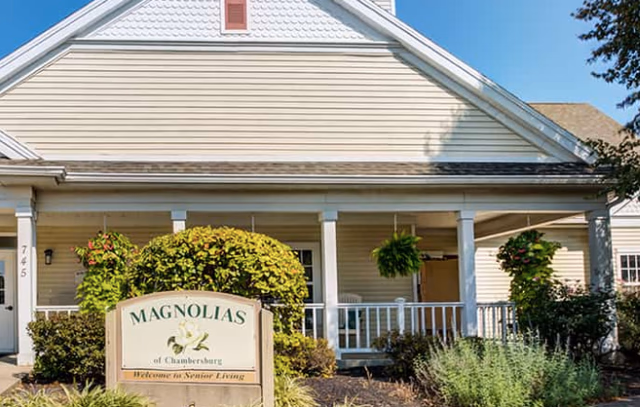 Exterior view of a single-story senior living facility building with beige siding and a white porch railing. There are hanging plants and bushes in front of the porch. A sign in the foreground reads 'Magnolias of Chambersburg Welcome to Senior Living.'