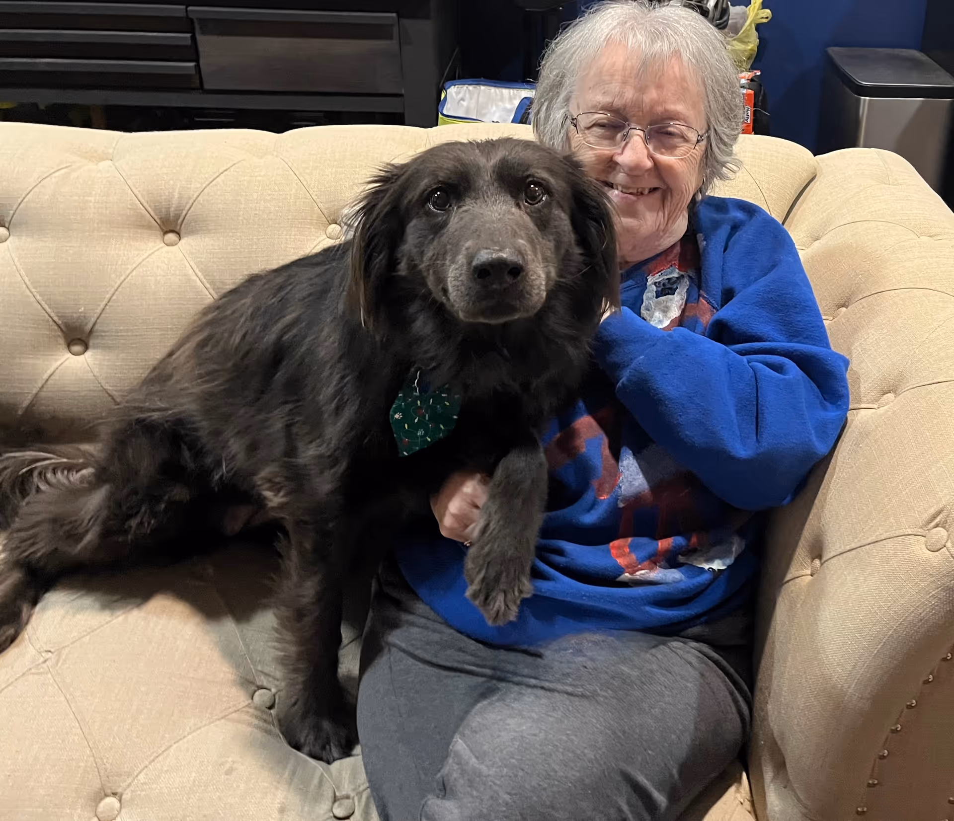 Elderly woman sits on a beige tufted couch with a black dog beside her.