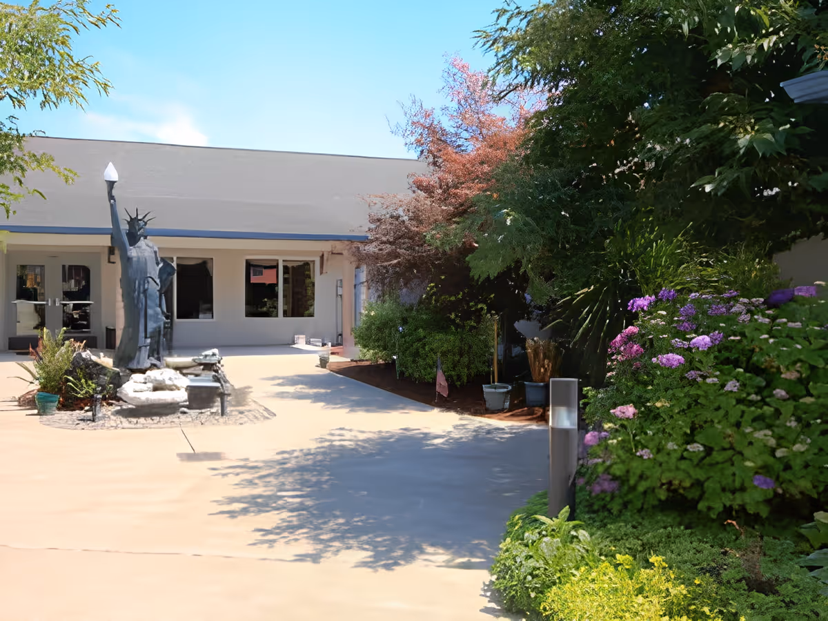 Outdoor courtyard area of a senior care facility with a statue resembling the Statue of Liberty, surrounded by greenery and colorful flowers, with a building entrance in the background under a clear blue sky.