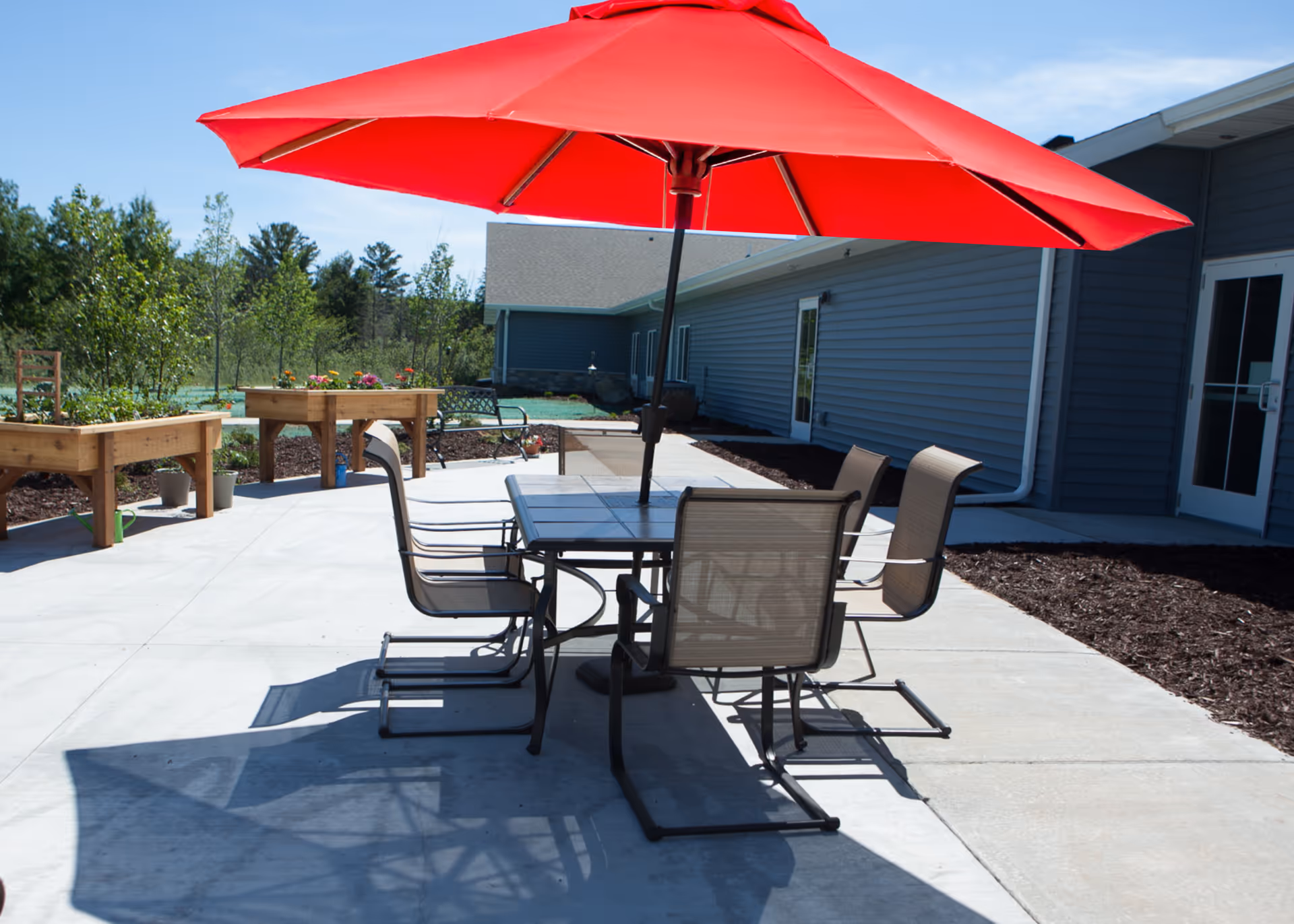 Outdoor patio with a table and chairs under a large red umbrella beside a single-story building.