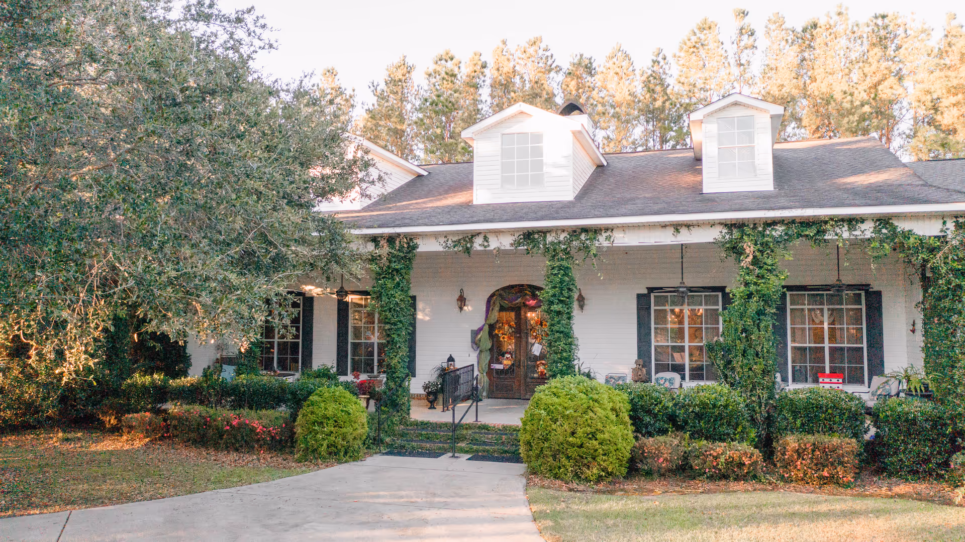 Front exterior view of a white building with a porch covered in green ivy and surrounded by bushes and trees. The building has three dormer windows on the roof and a pathway leading to the entrance door decorated with wreaths and flowers.