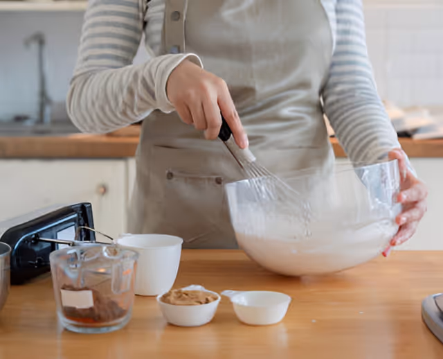 Person in an apron whisking batter in a kitchen with bowls on the counter.