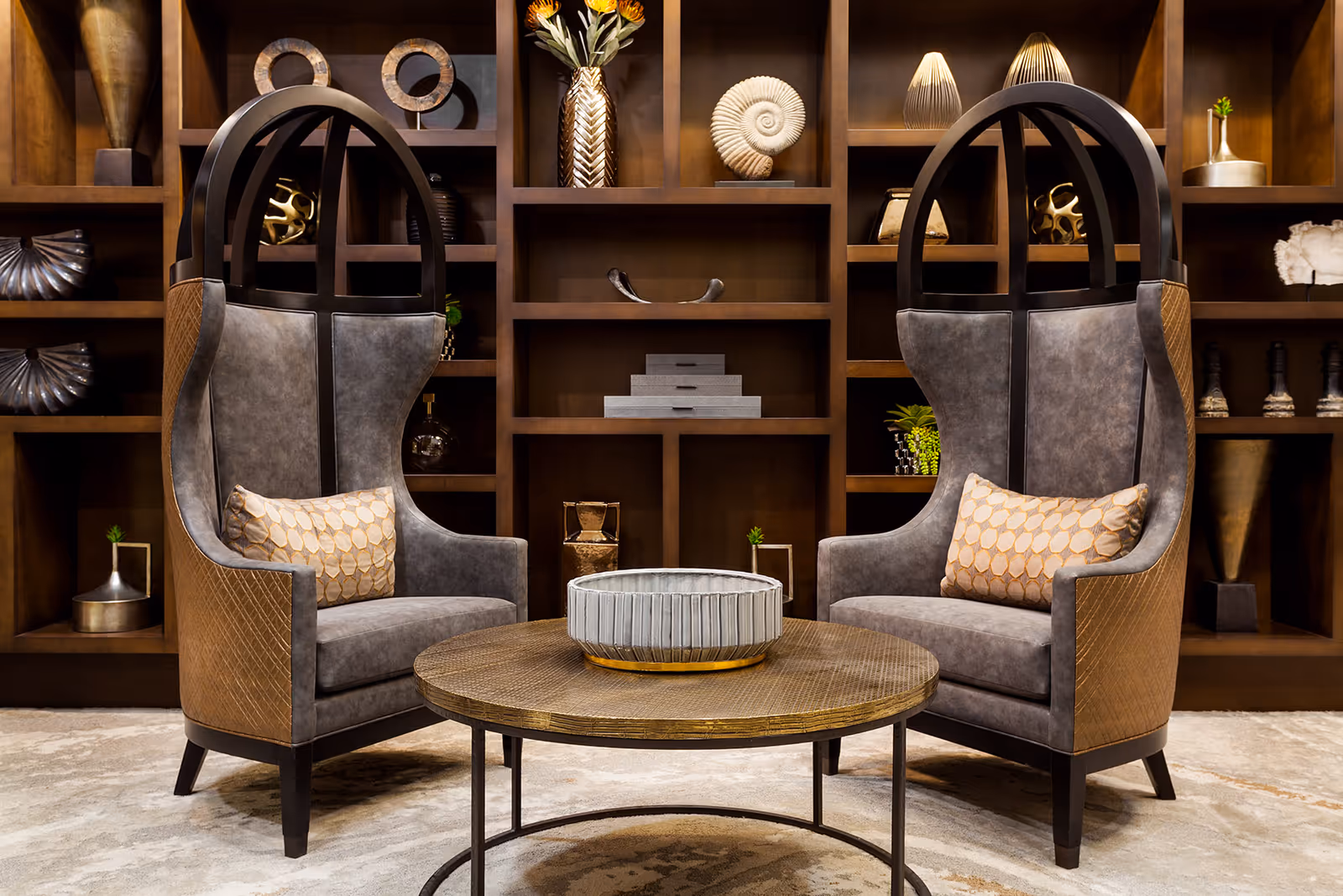 Two high-backed, modern armchairs with gray upholstery and brown quilted sides, each with a decorative pillow, positioned around a round wooden coffee table with a large decorative bowl on top. Behind the chairs is a large wooden shelving unit filled with various decorative objects including vases, sculptures, and plants.