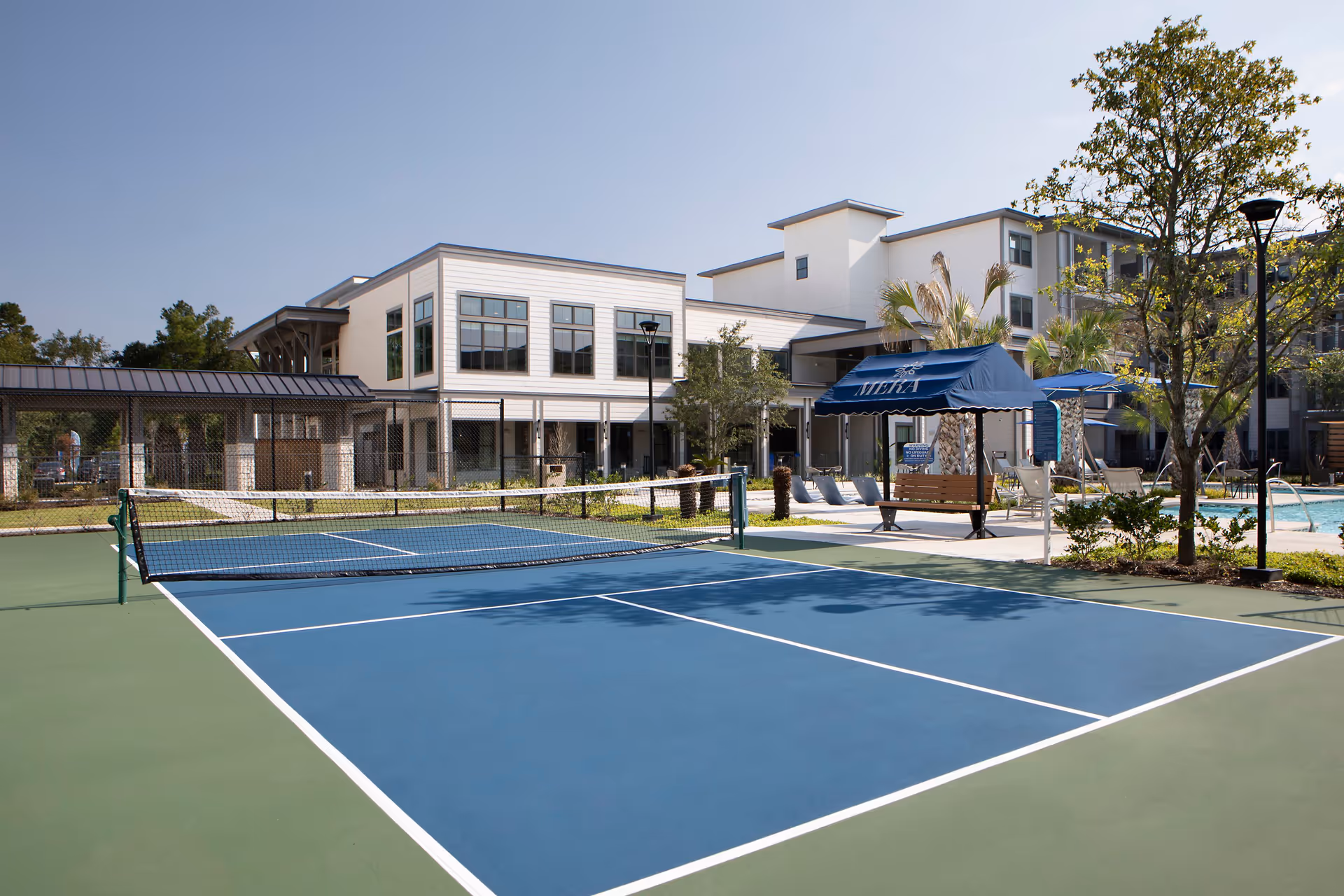 Outdoor tennis court with a net in the foreground, surrounded by a fence. In the background, there is a modern multi-story building with large windows, a shaded seating area with a blue canopy labeled 'MERA', palm trees, and a swimming pool area with lounge chairs. The sky is clear and blue.