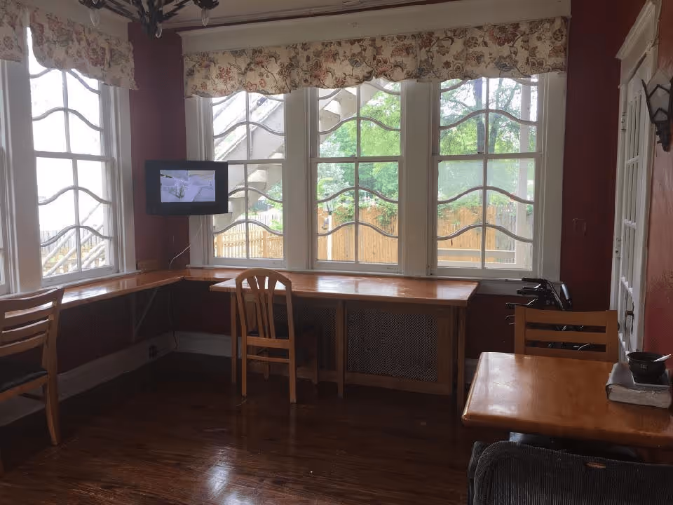 Sunlit common room with wooden chairs and tables along large decorative windows and a small wall-mounted TV.