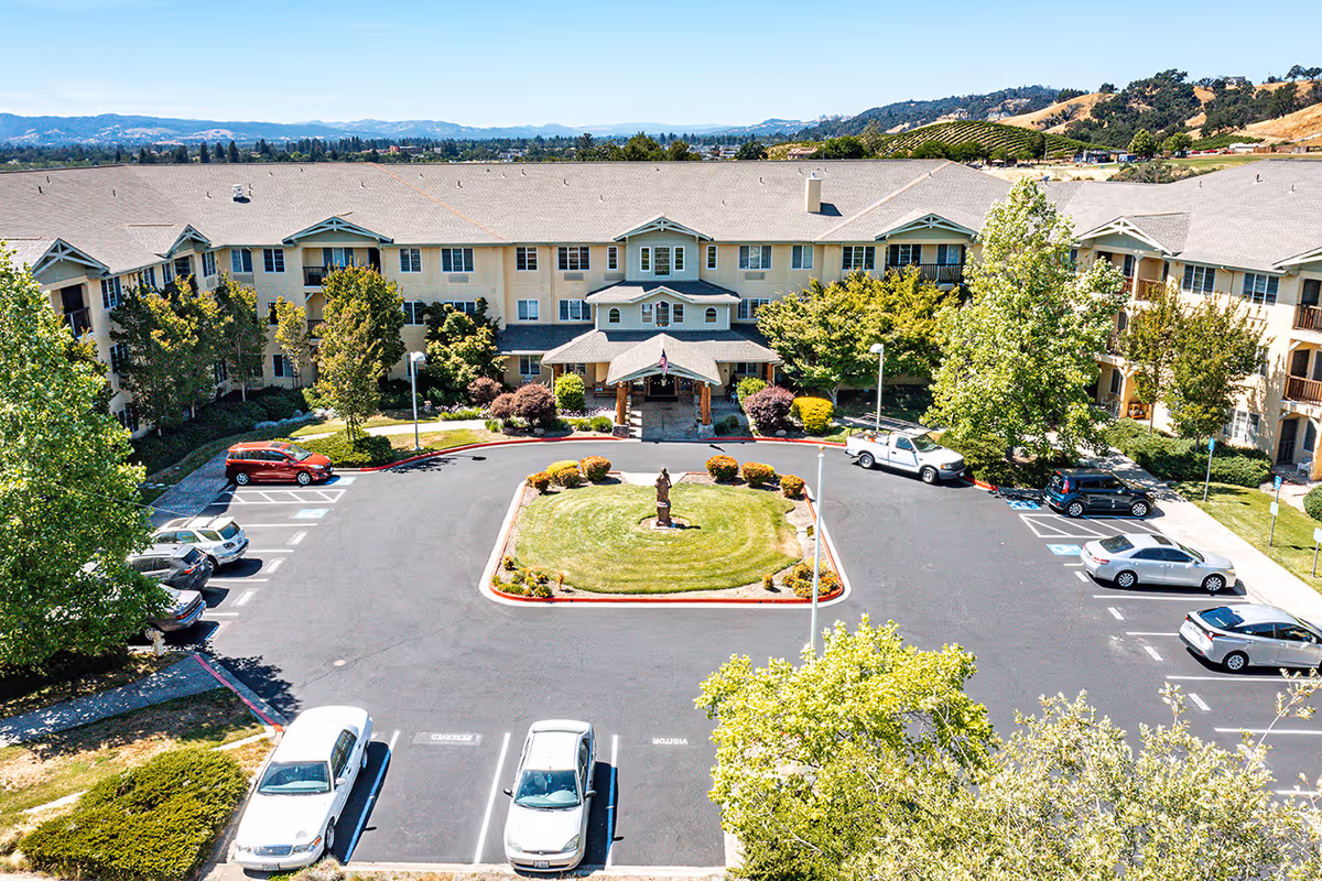 Aerial view of a three-story senior living building with a central entrance, circular landscaped driveway and parked cars in the lot.