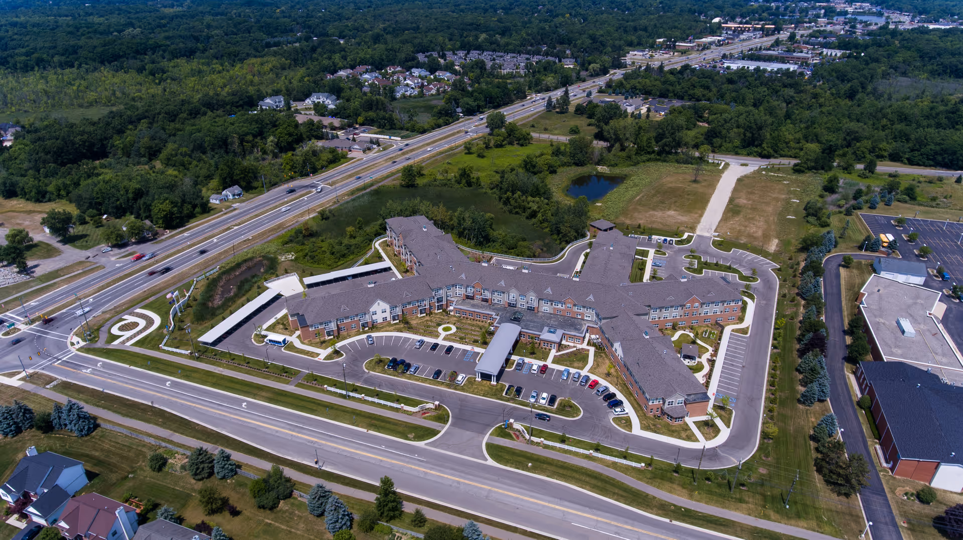 Aerial view of Pomeroy Living Orion Assisted Living facility showing a large, multi-wing building surrounded by parking lots, roads, and green spaces with trees and a small pond nearby.