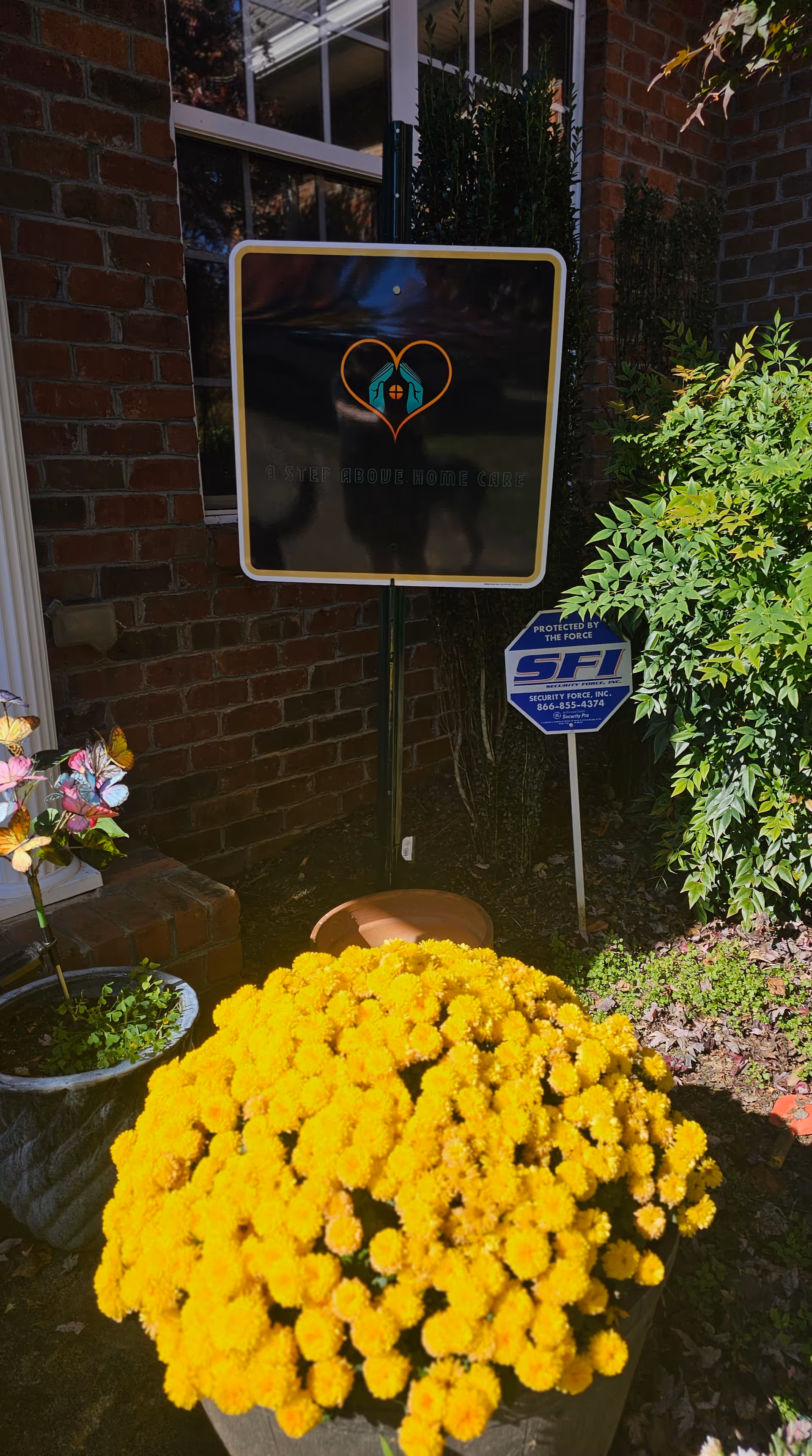 Front entrance area with a signboard, brick wall, and bright yellow potted chrysanthemums.