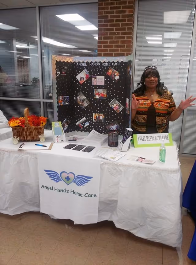 A woman stands beside a display table for Angel Hands Home Care in an indoor booth.