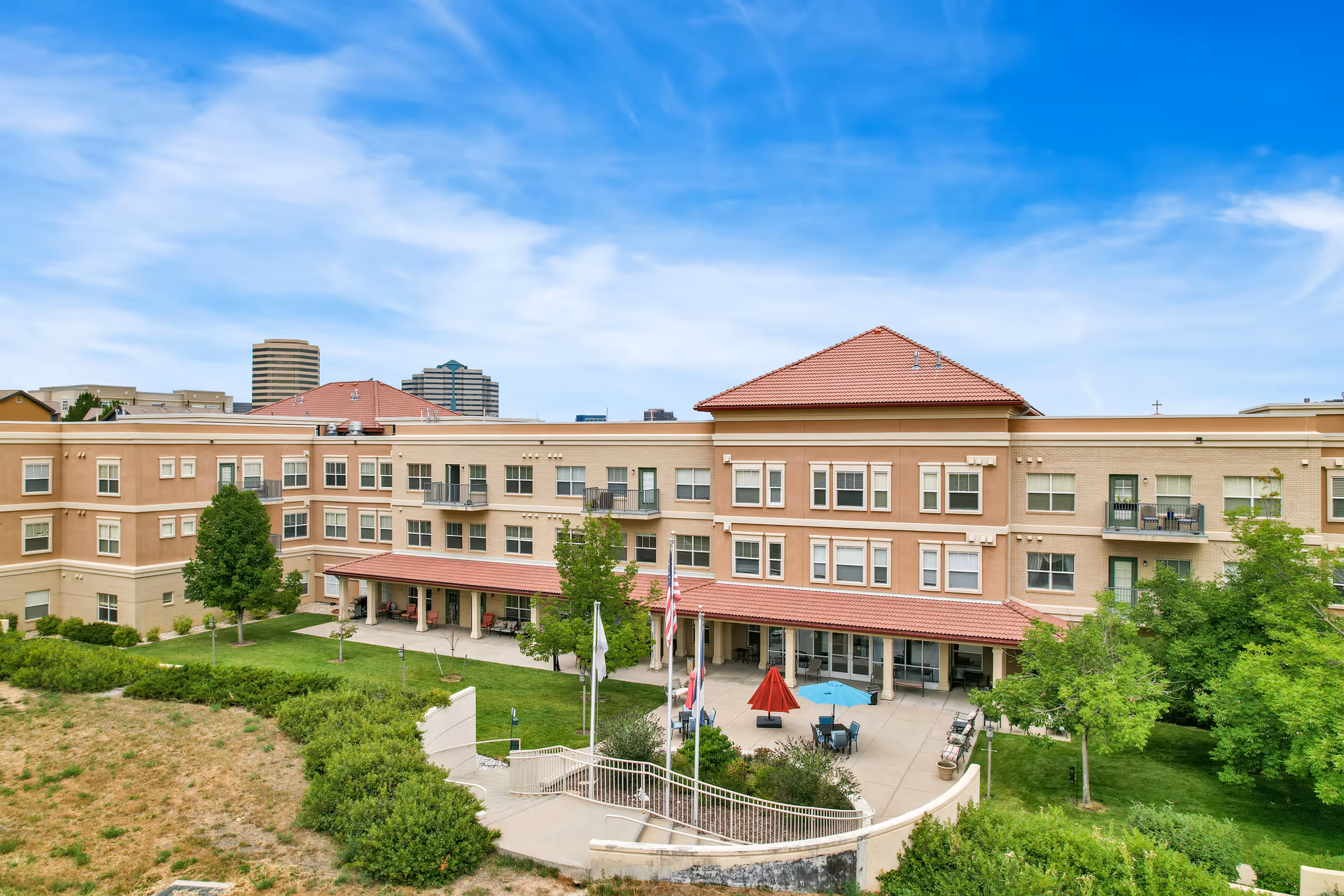 Exterior view of The Inn at Greenwood Village, a multi-story senior living facility with beige and light brown walls, red tiled roofs, balconies, and a patio area with tables and umbrellas surrounded by green trees and shrubs under a blue sky with scattered clouds.
