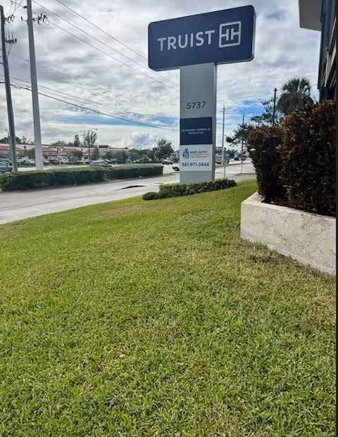 Blue TRUST sign on a grassy roadside area near a hedge and building edge
