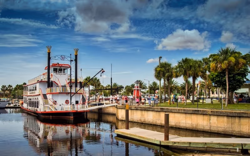 Large paddleboat docked beside a waterfront park with palm trees and people.