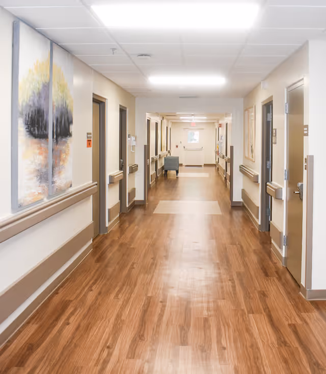 Well-lit interior hallway of a care facility with wood-look flooring, handrails and doors to rooms on both sides.