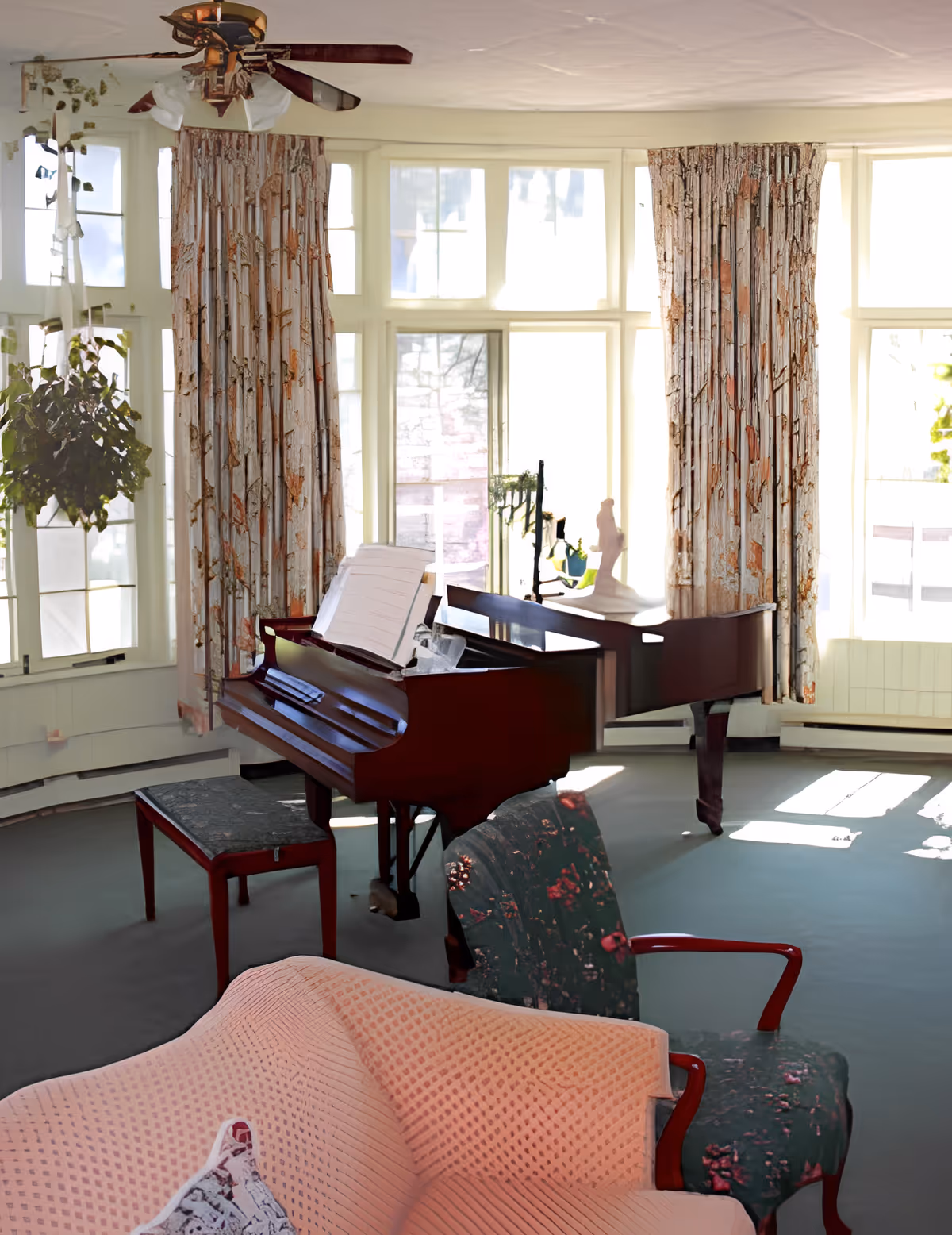 Sunlit sitting room with a grand piano in front of large bay windows, upholstered chairs and a sofa.