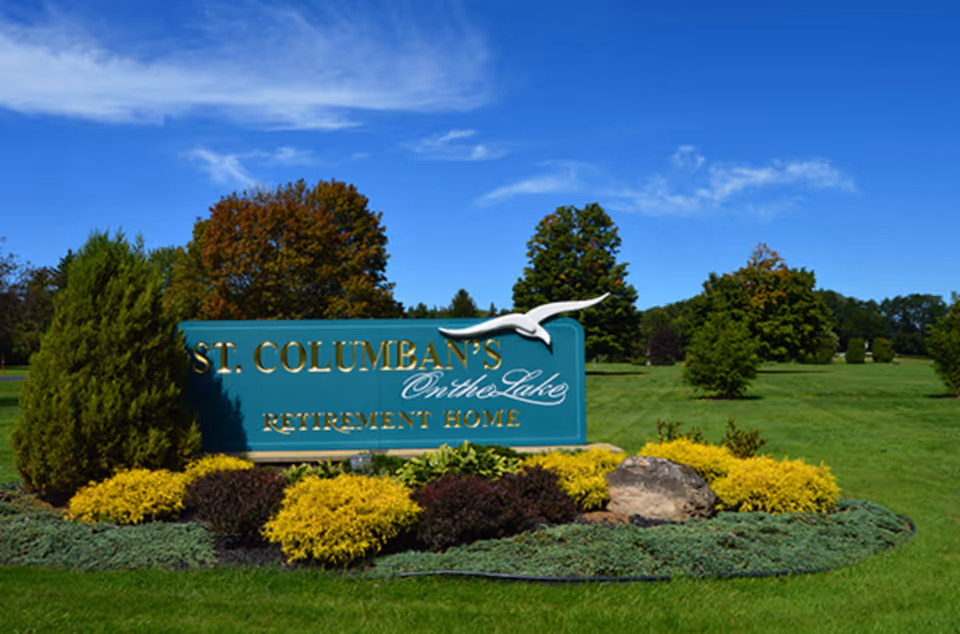 A large green sign with gold lettering that reads 'St. Columban's on the Lake Retirement Home' surrounded by well-maintained landscaping including bushes, small trees, and rocks under a clear blue sky.