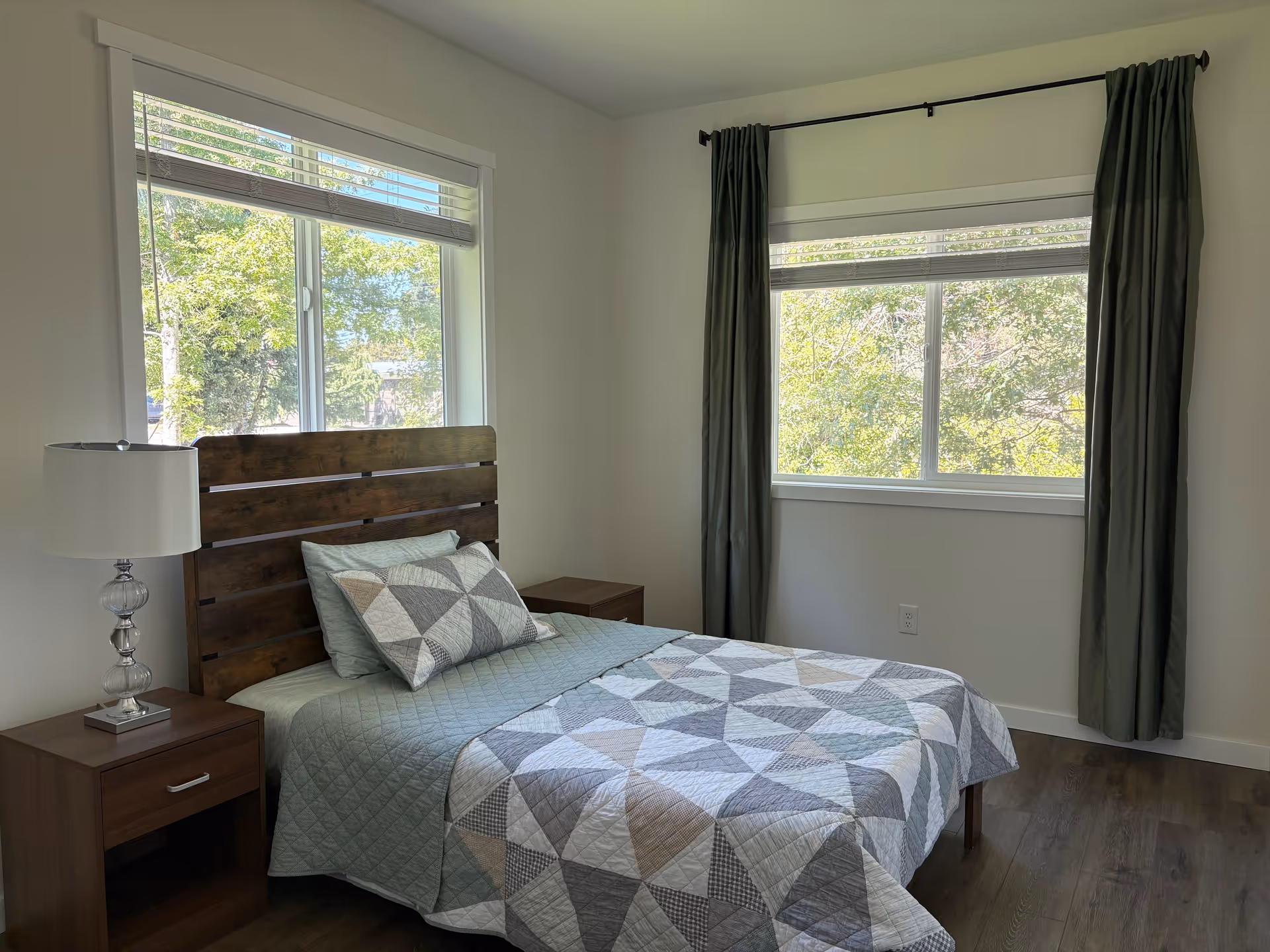 A tidy bedroom with a wooden headboard, quilted bedspread, and two windows with curtains.