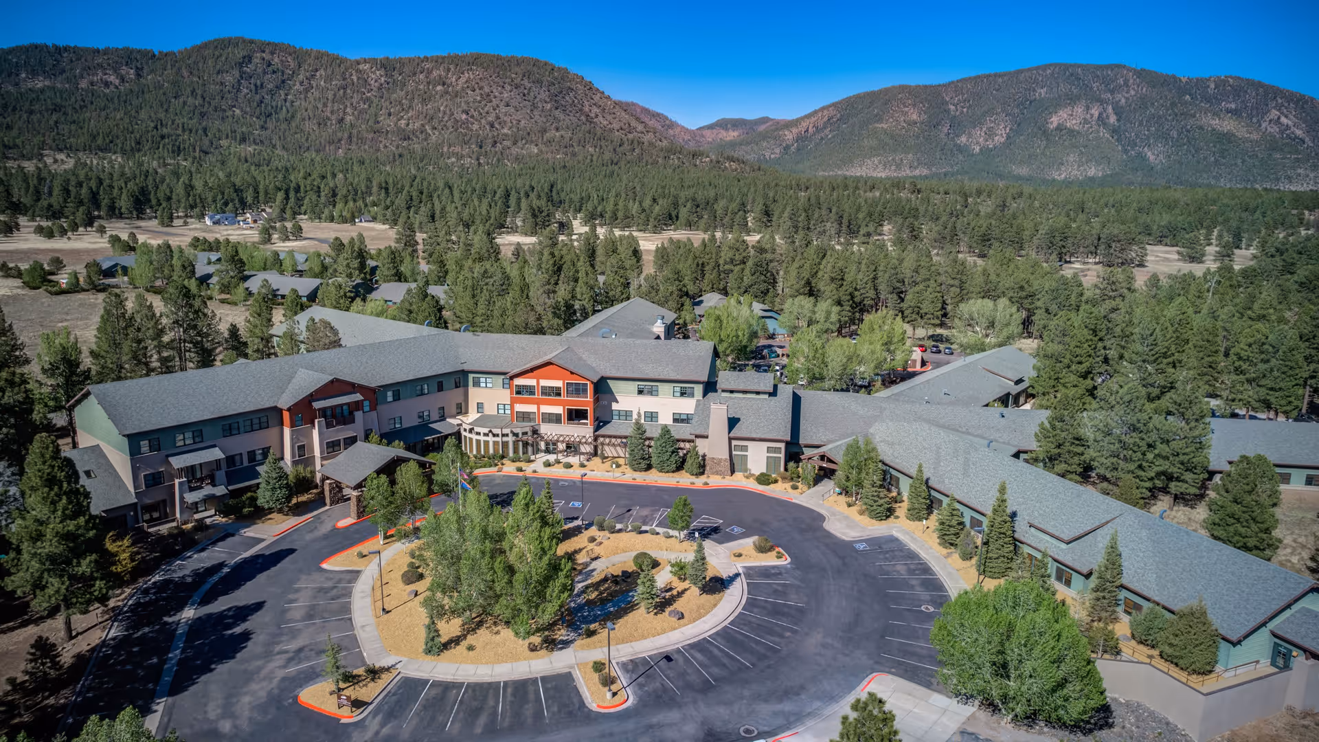 Aerial view of The Peaks, A Senior Living Community, showing a large building complex surrounded by trees and mountains in the background. The building has multiple wings with gray roofs and a circular driveway with parking spaces in front. The area is lush with greenery and natural landscape.