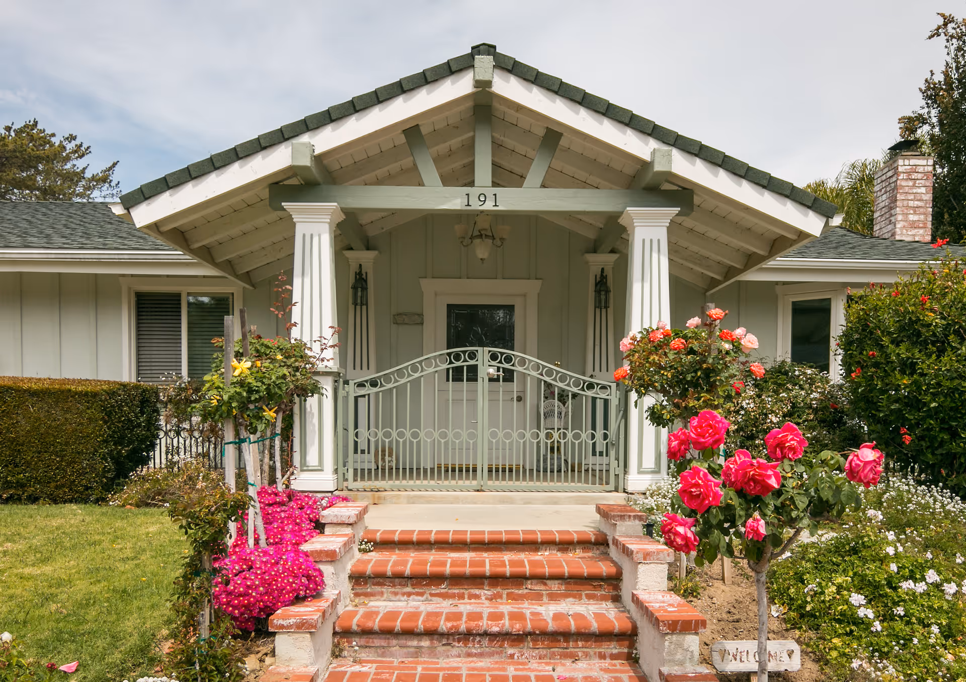 Front entrance of a single-story house with a covered porch supported by white columns. The house number 191 is displayed above the porch. There is a green metal gate at the top of brick steps leading to the front door. The garden around the entrance is well-maintained with vibrant pink and red flowers and green shrubs.