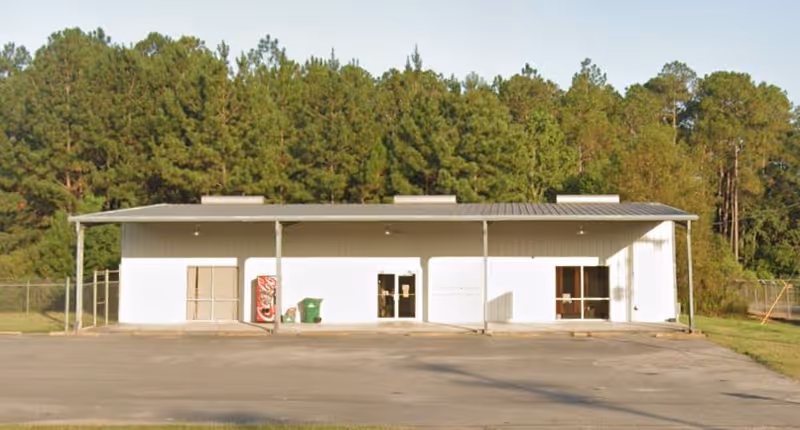 Front view of a single-story white building with a metal roof overhang and trees behind.