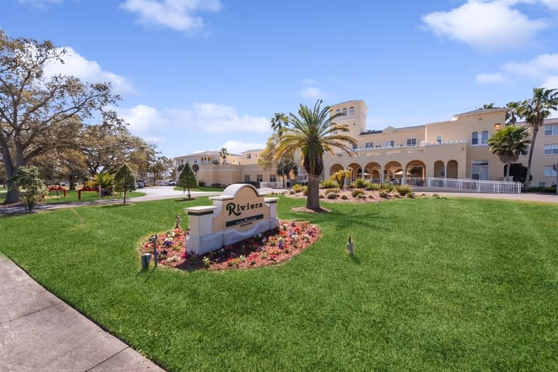 Front exterior view of Riviera Senior Living facility with a manicured lawn, palm trees, and a sign displaying the facility's name. The building has a beige facade with arches and multiple windows under a partly cloudy blue sky.