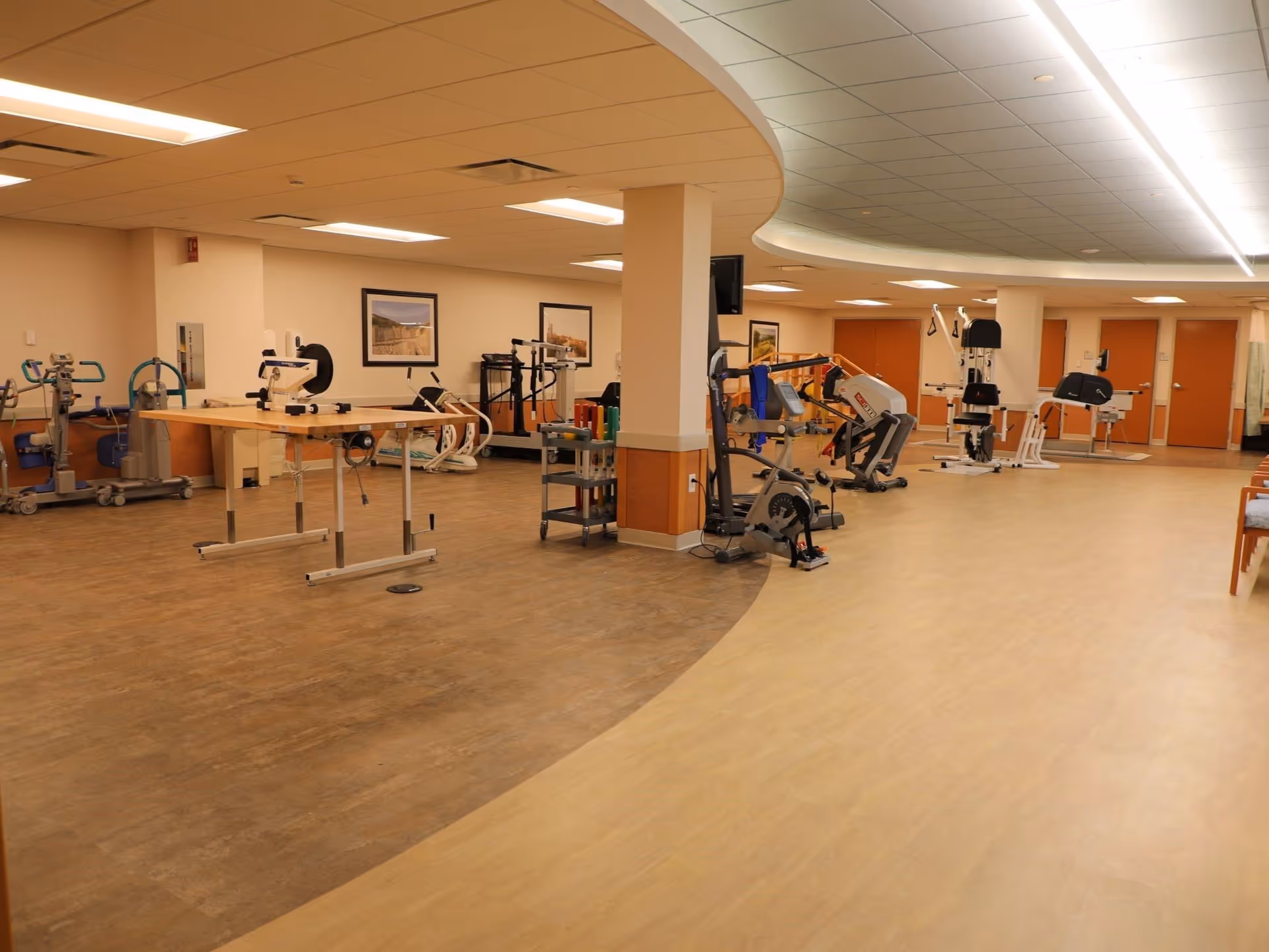 A spacious rehabilitation room with various exercise and therapy equipment including stationary bikes, parallel bars, and other physical therapy machines. The room has a curved ceiling design, wood and light-colored flooring, and framed pictures on the walls.