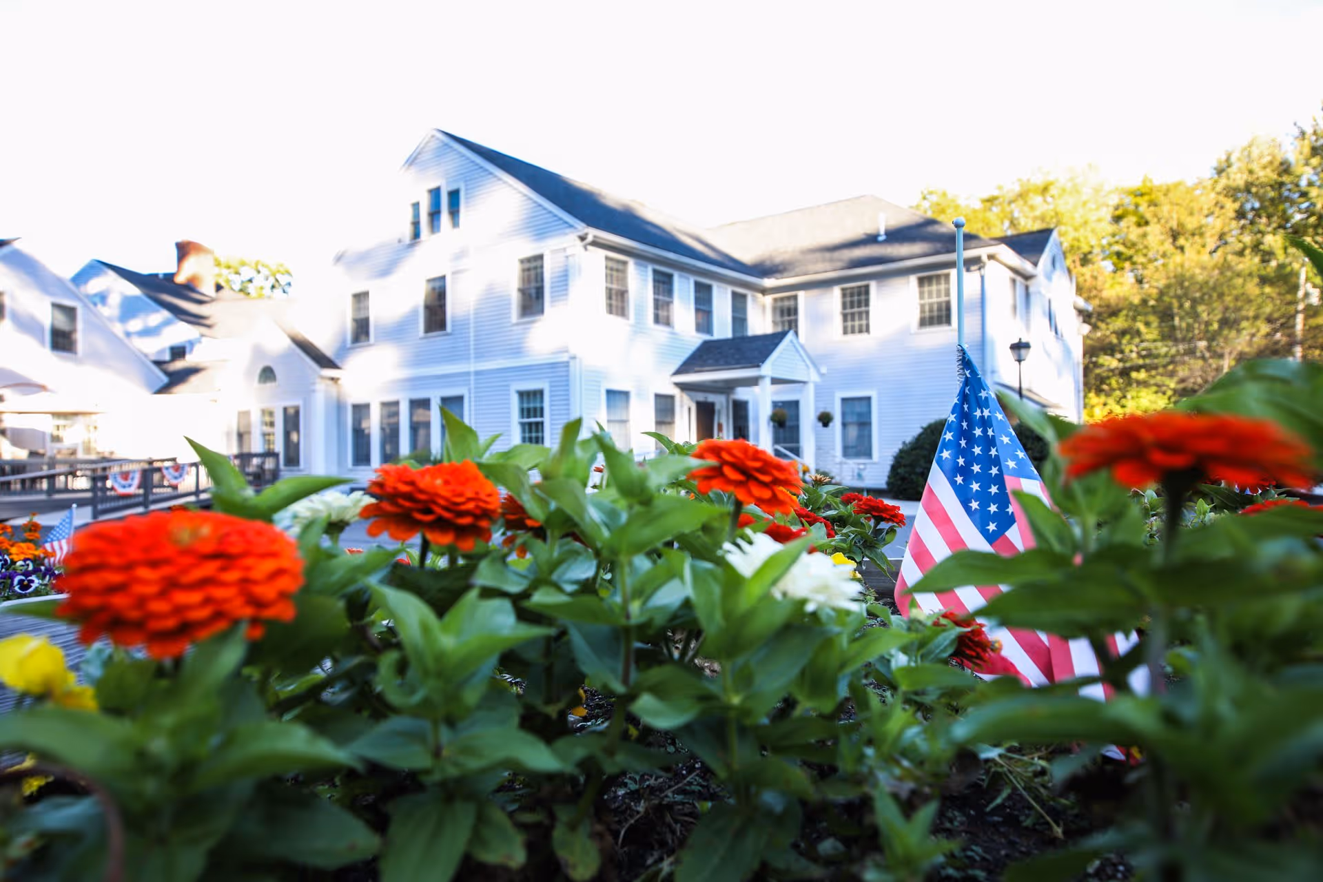 A white multi-story building with many windows, partially obscured by a garden bed filled with vibrant red and white flowers and an American flag. Trees and greenery are visible in the background under a clear sky.