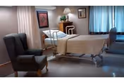 Hospital-style bed in a furnished bedroom with chairs and window drapes.