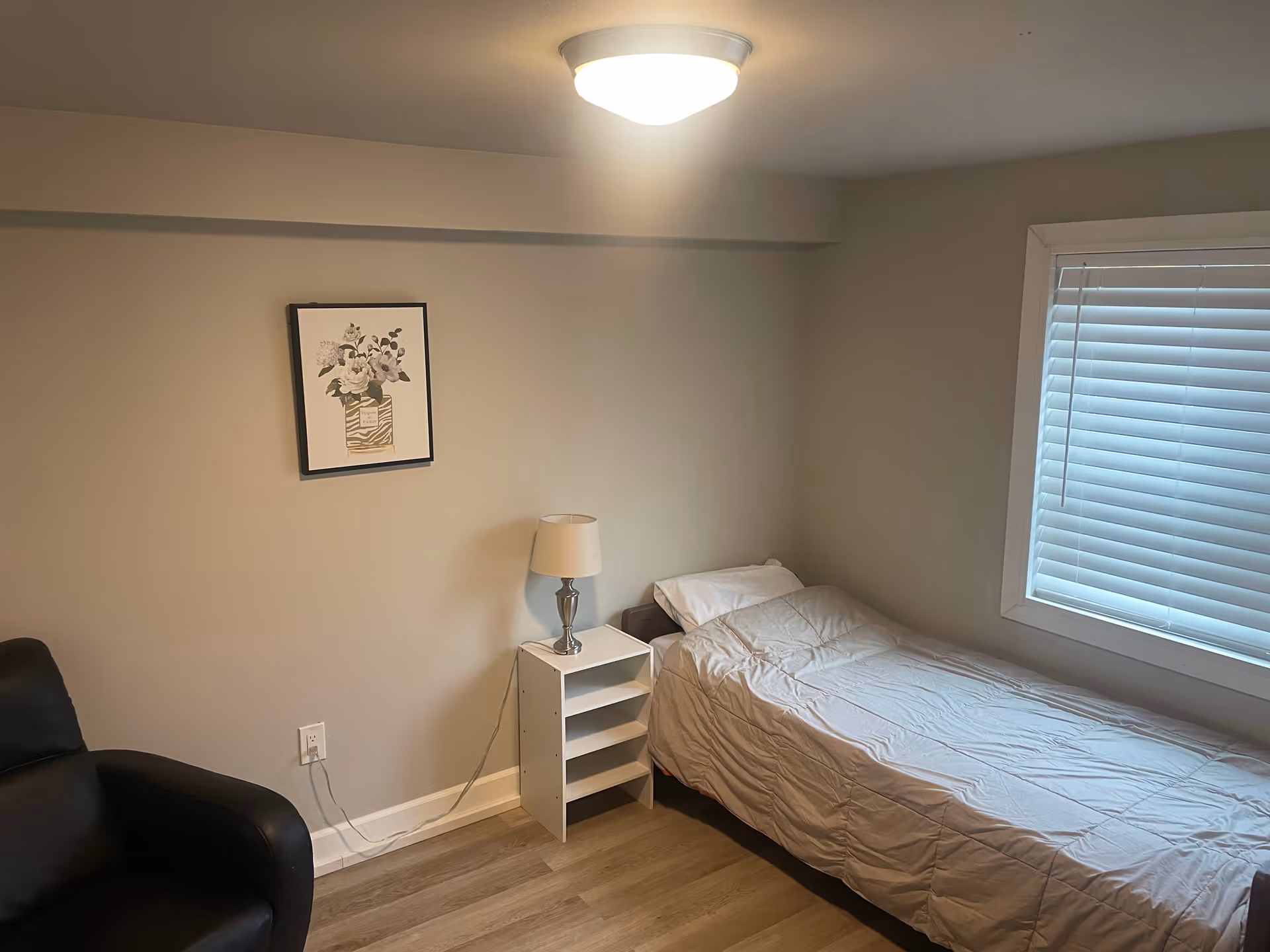 Small bedroom with single bed, nightstand, lamp, framed floral art, and window blinds.
