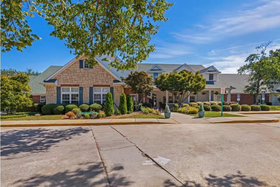 Exterior view of a senior living facility building with a stone and brick facade, surrounded by well-maintained landscaping including bushes, trees, and potted plants. The sky is clear and blue, and there is a paved driveway in front with a white directional arrow painted on it.