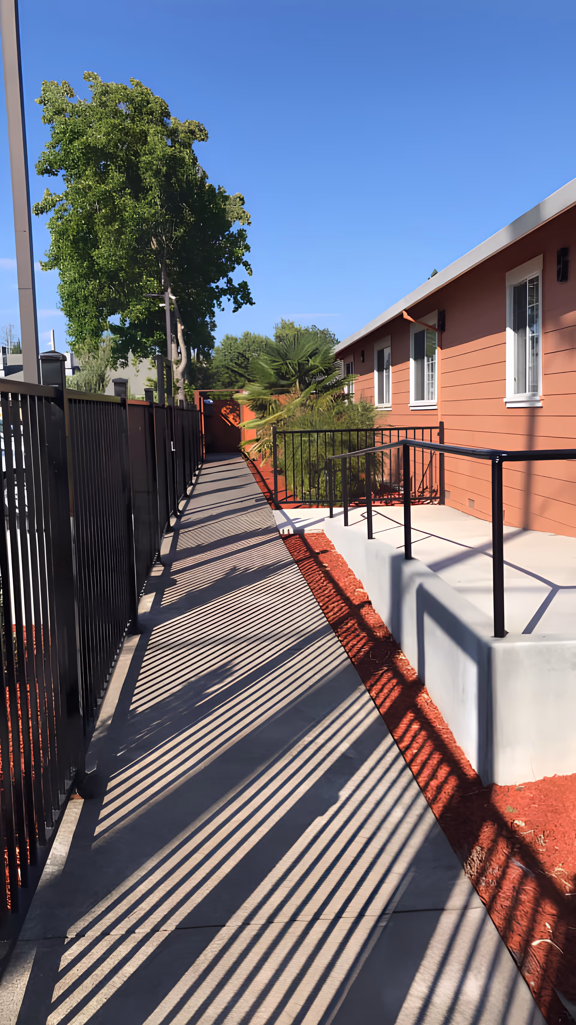 Outdoor walkway beside a red single-story building with metal railings casting striped shadows under a clear blue sky.