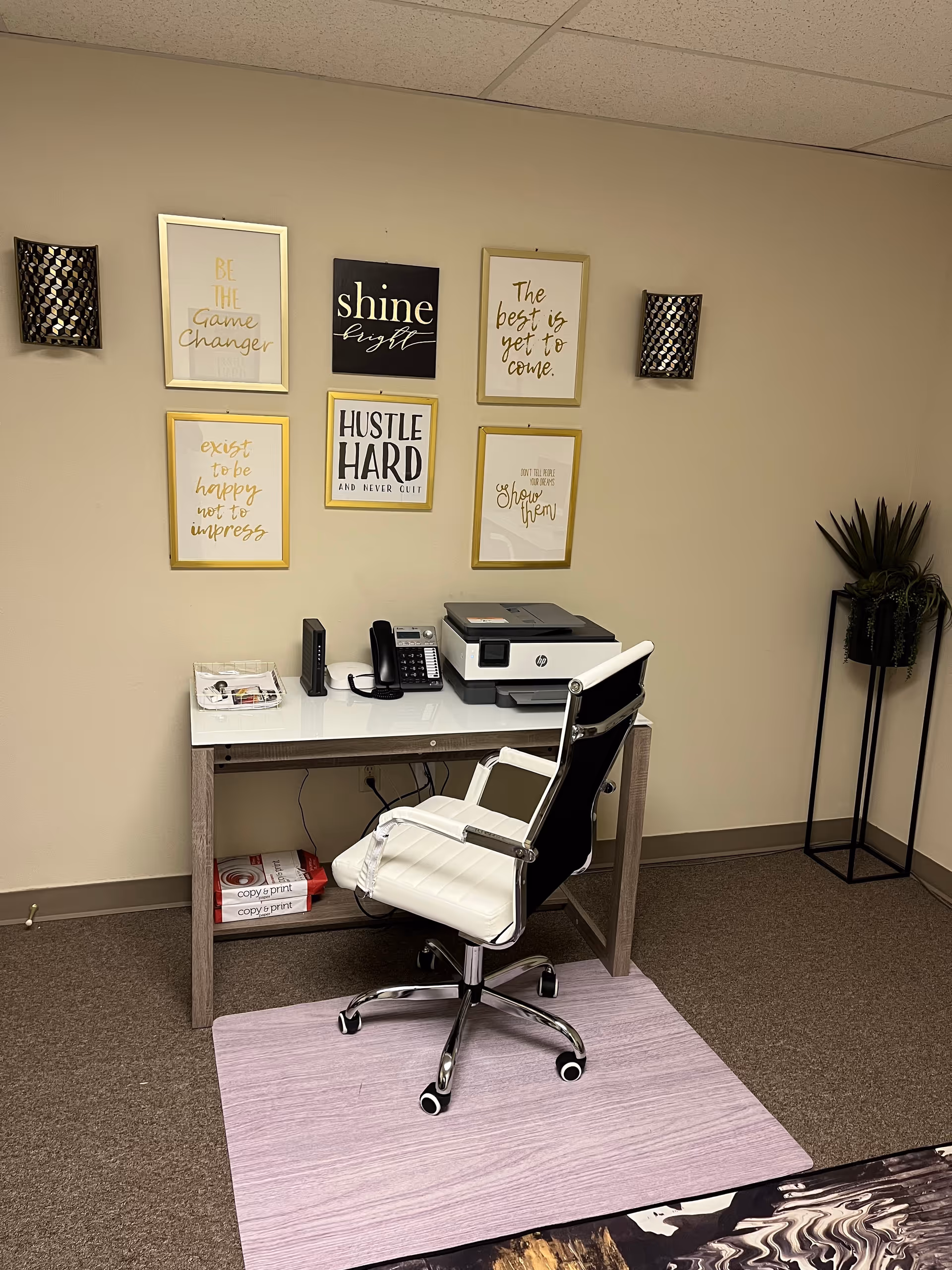 A white office desk with printer, phone, and framed quotes on a beige wall.