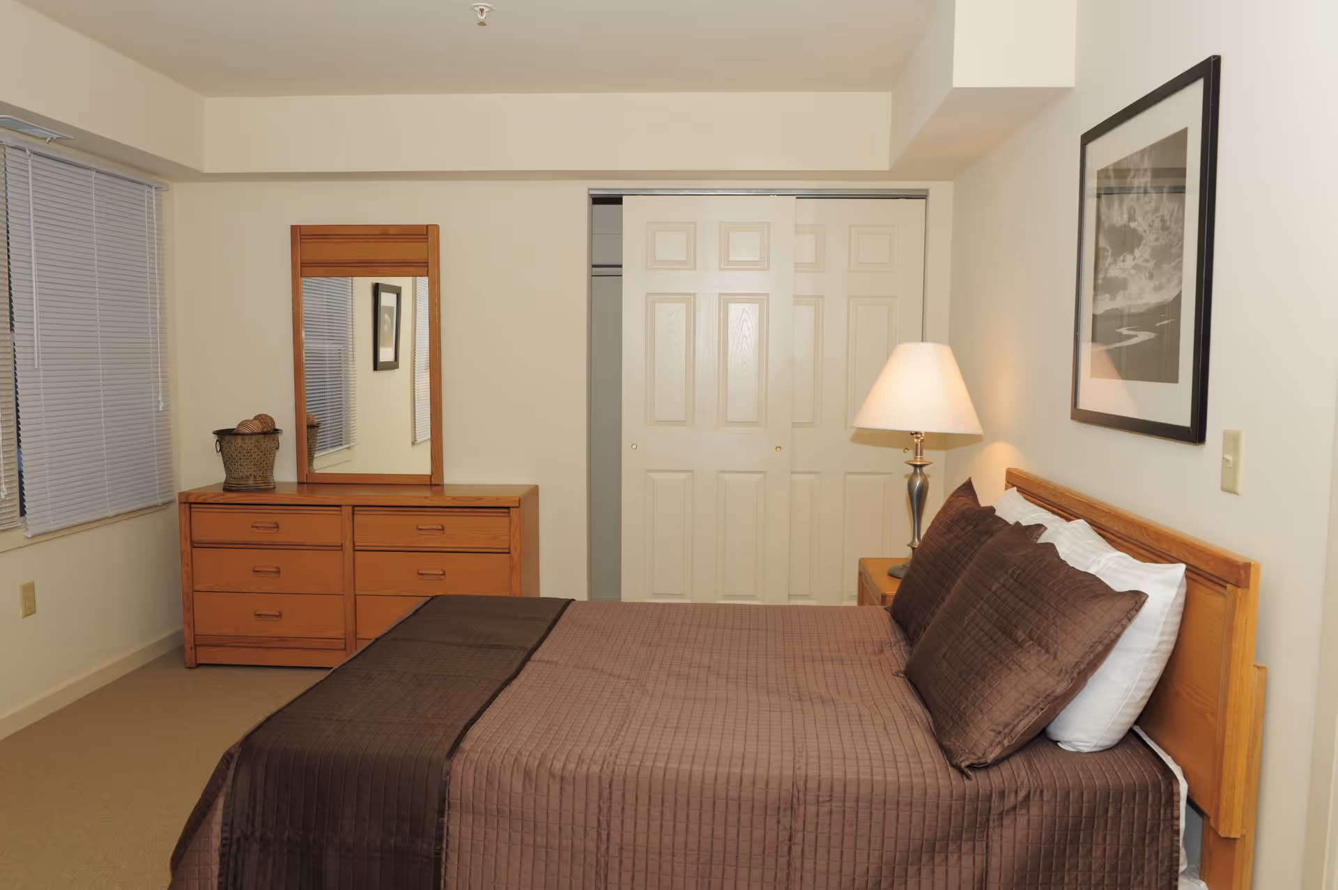 A bedroom with a neatly made bed featuring brown bedding and pillows. To the right of the bed is a wooden nightstand with a lamp. On the wall above the bed is a framed black and white picture. Across from the bed is a wooden dresser with a mirror and a decorative basket on top. There is a window with closed blinds on the left wall and a closet with sliding doors in the center background.