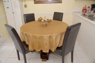 Round dining table covered with a gold tablecloth and a decorative bowl, surrounded by four dark upholstered chairs in a kitchen/dining area.