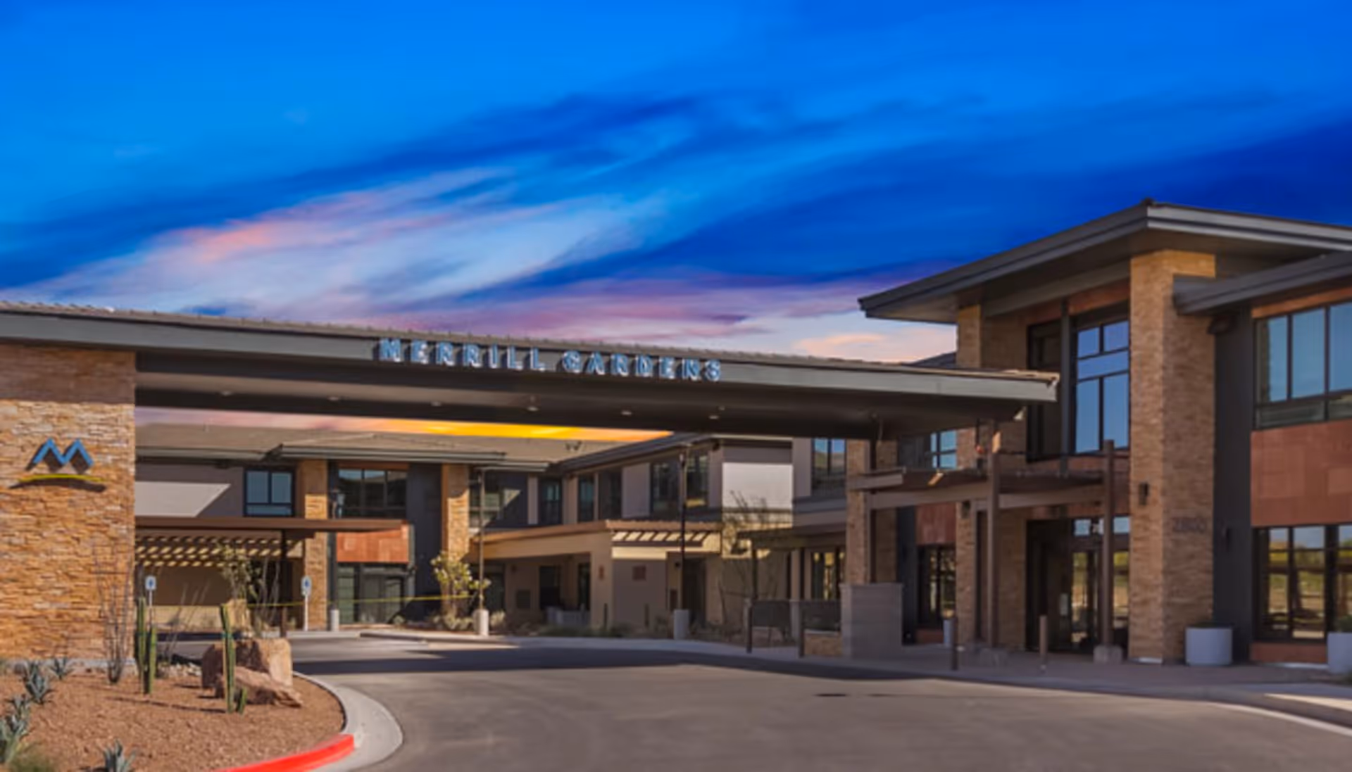 Exterior view of Merrill Gardens at Anthem building during sunset with a clear sky showing the entrance driveway and modern architectural design featuring large windows and stone accents.