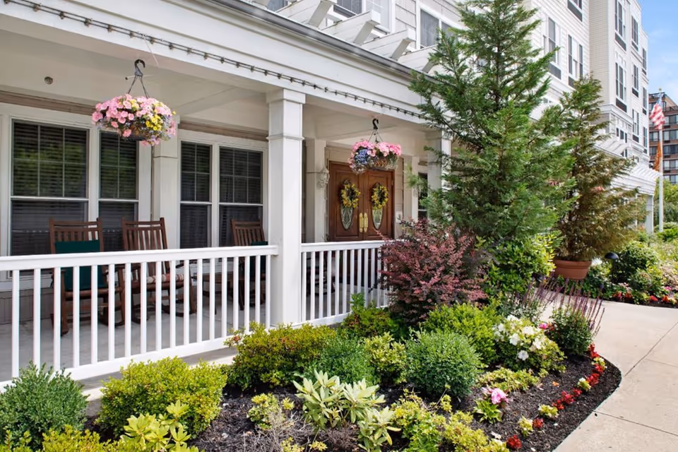 Front porch of Sunrise of Staten Island senior living facility with chairs and hanging baskets.