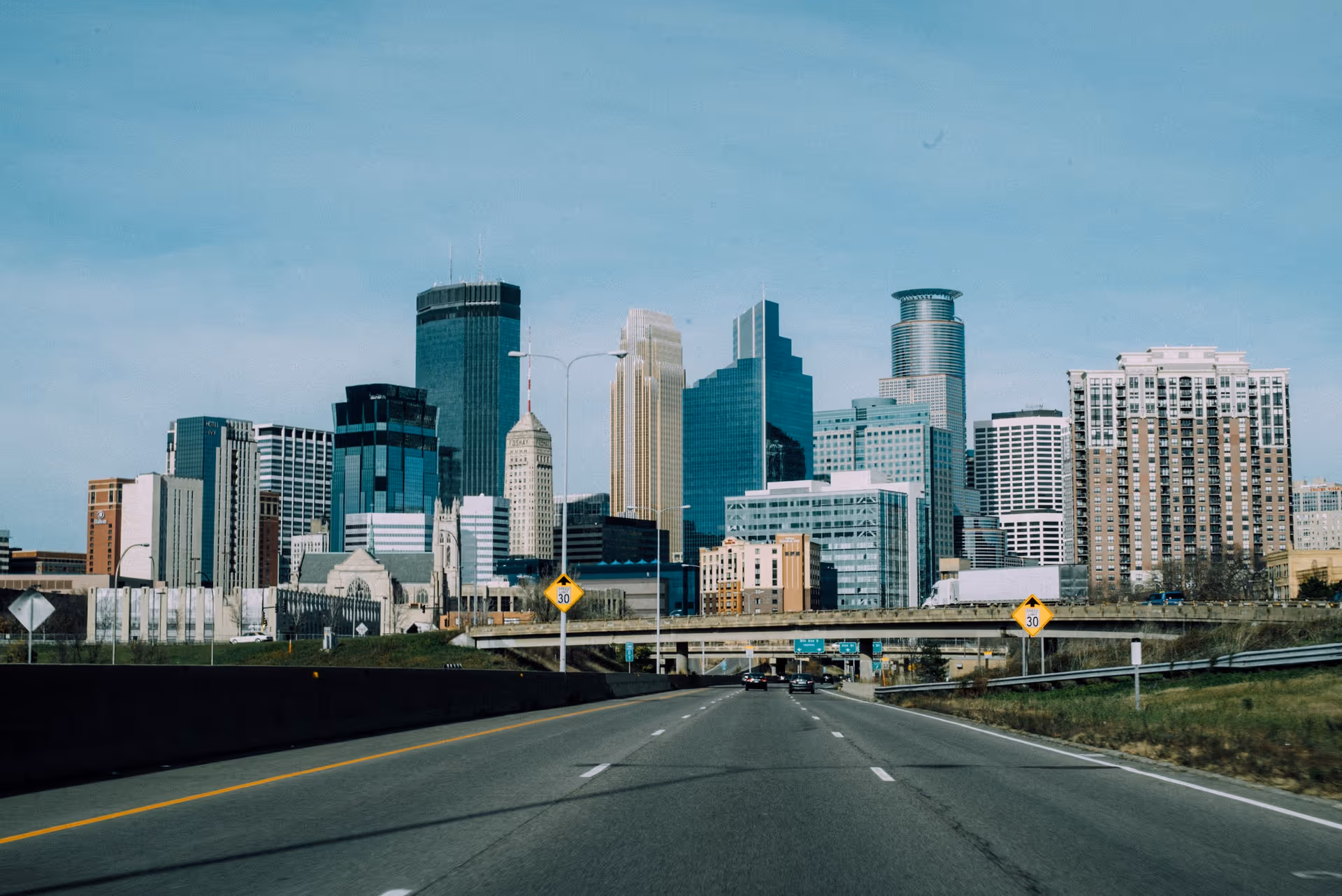A highway leads toward a downtown skyline with tall modern buildings under a clear blue sky.