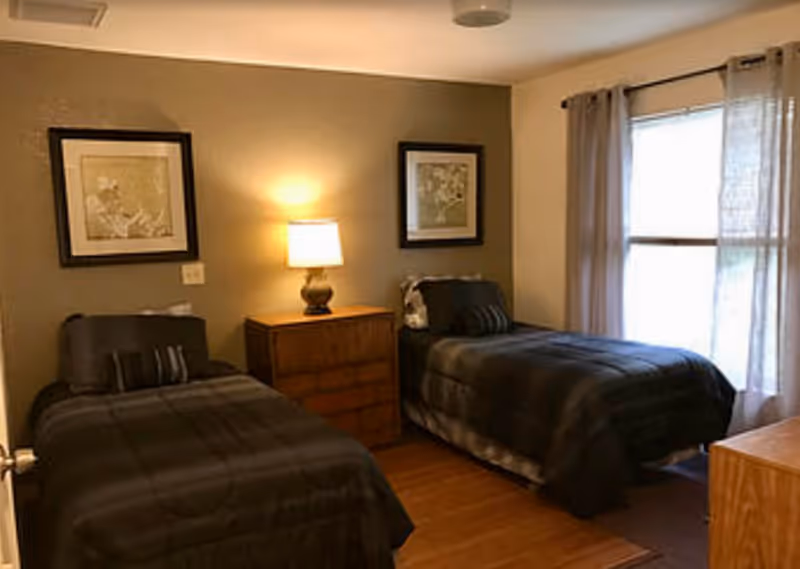 Two twin beds flank a dresser with a lamp, framed art, and window curtains.