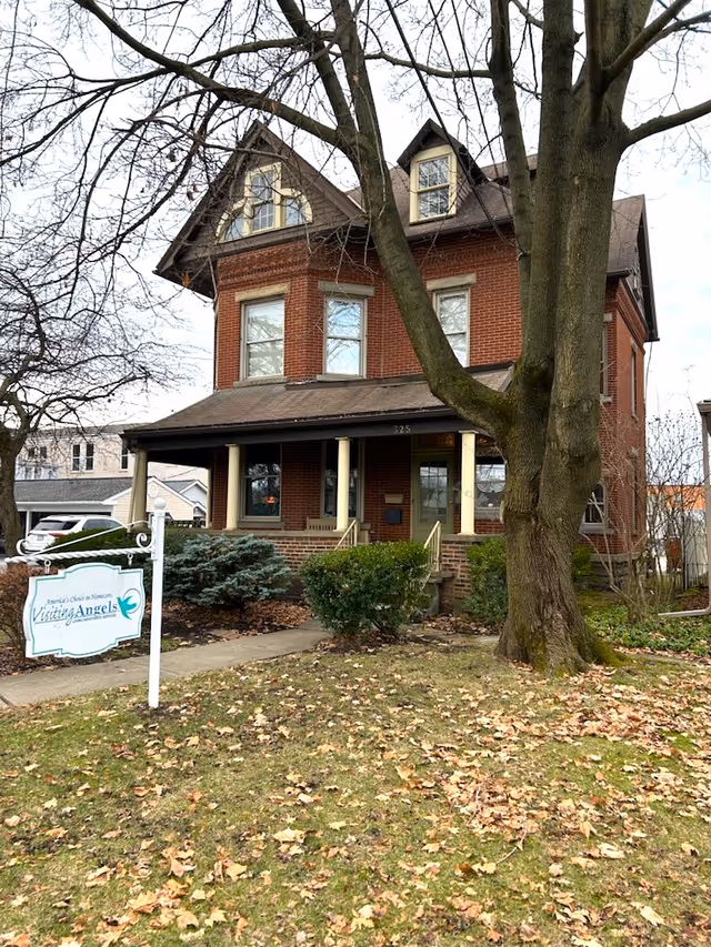 This brick building front features a porch, large trees, and a Visiting Angels sign.