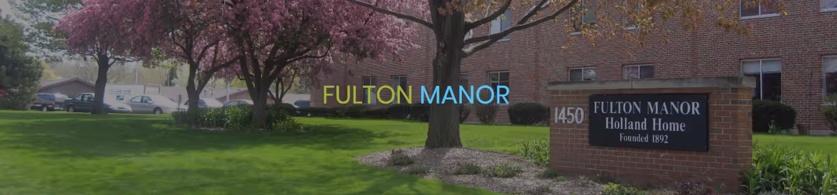 Exterior view of Fulton Manor, showing a brick building with several windows, a green lawn with blooming pink trees, and a brick sign that reads '1450 Fulton Manor Holland Home Founded 1892'.