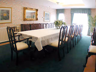 Formal dining room with a long rectangular table covered by a white cloth surrounded by wooden chairs, a sideboard, framed artwork, and windows with drapes.