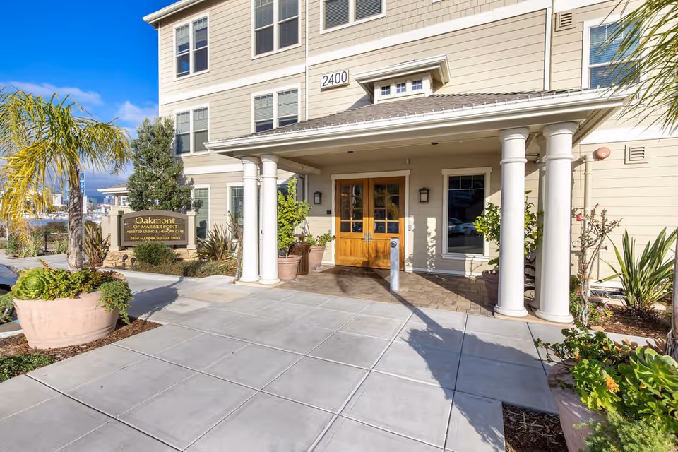 Entrance of Oakmont of Mariner Point assisted living and memory care facility with a covered porch supported by white columns, wooden double doors, potted plants, and a sign displaying the facility name and address.