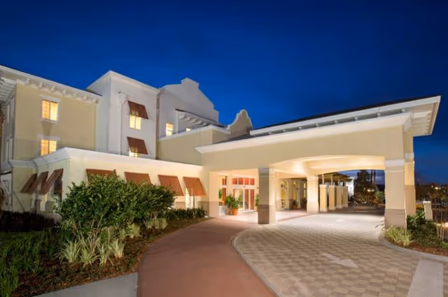 Exterior view of a senior living facility building at dusk with illuminated windows and entrance. The building has beige walls, red window awnings, and a covered driveway with an arrow on the pavement. There are landscaped bushes and plants along the walkway.